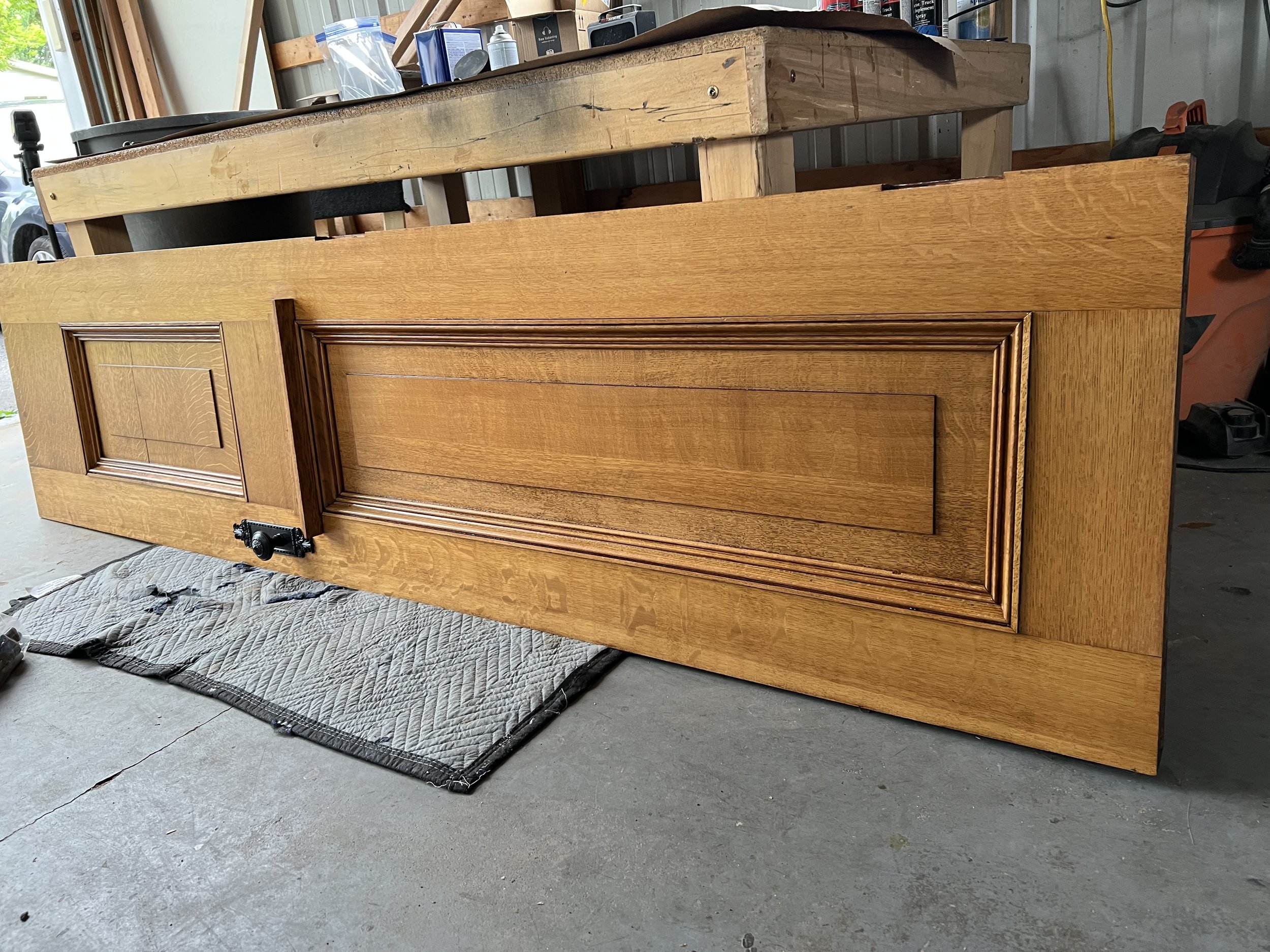 A wooden door lying on its side on a concrete garage floor, with a quilted mat underneath. The cabinet has two paneled doors and is in a workshop or garage, with tools, cans, and shelves visible in the background.