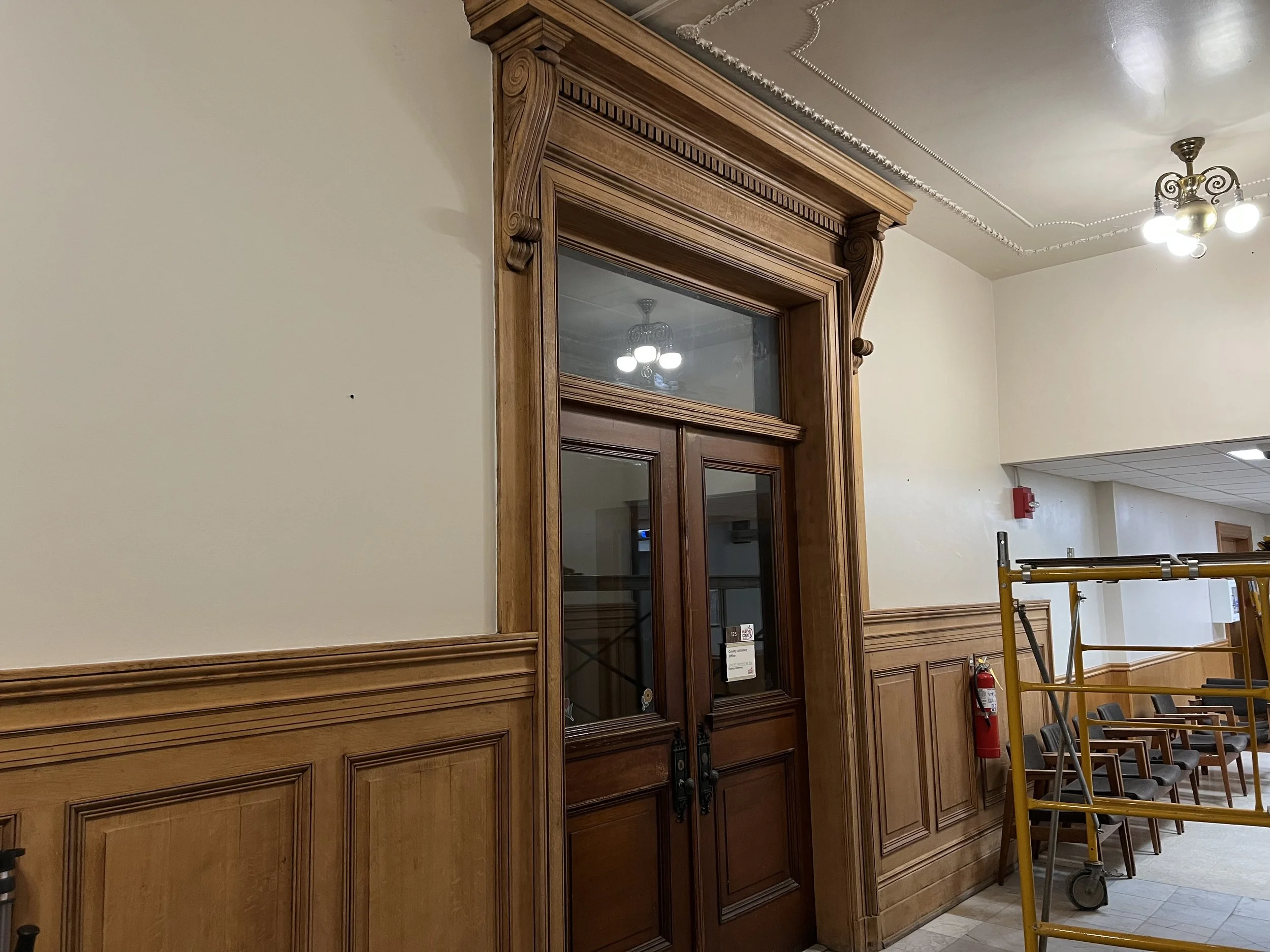 Interior of a building with decorative wooden molding around a doorway, a ceiling with ornate crown molding, and a chandelier with multiple lights.
