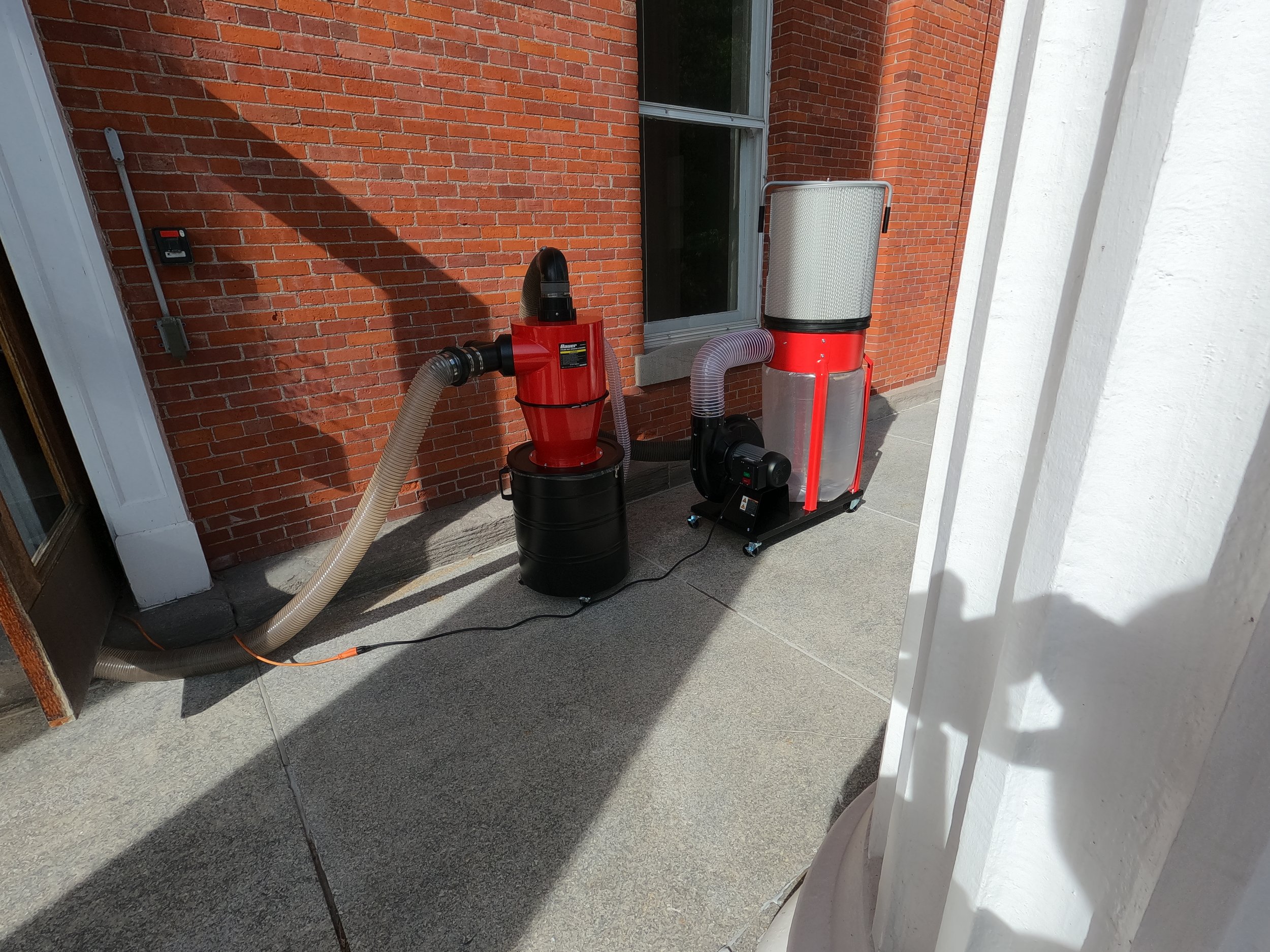 Outdoor scene with equipment attached to a brick wall, including a vacuum or filtration system with hoses, on a concrete patio next to a white wall and a sliding glass door.