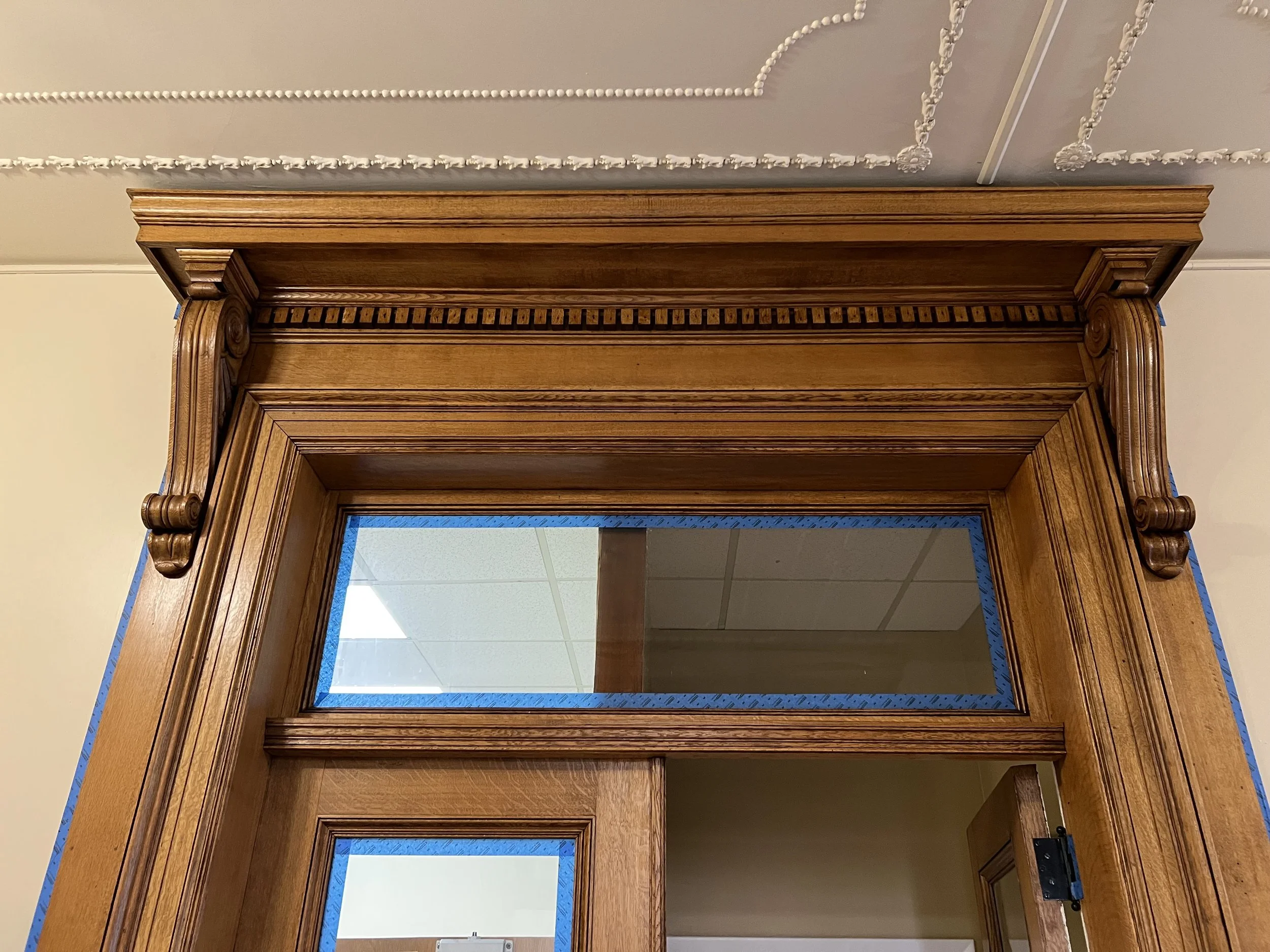 The image shows a wooden doorway with ornate molding and a mirror attached to the top part of the frame. The ceiling is decorated with intricate plasterwork, and there is some blue painter's tape around the edges of the doorway. Through the mirror, a ceiling with white ceiling tiles and a lighting fixture is visible.