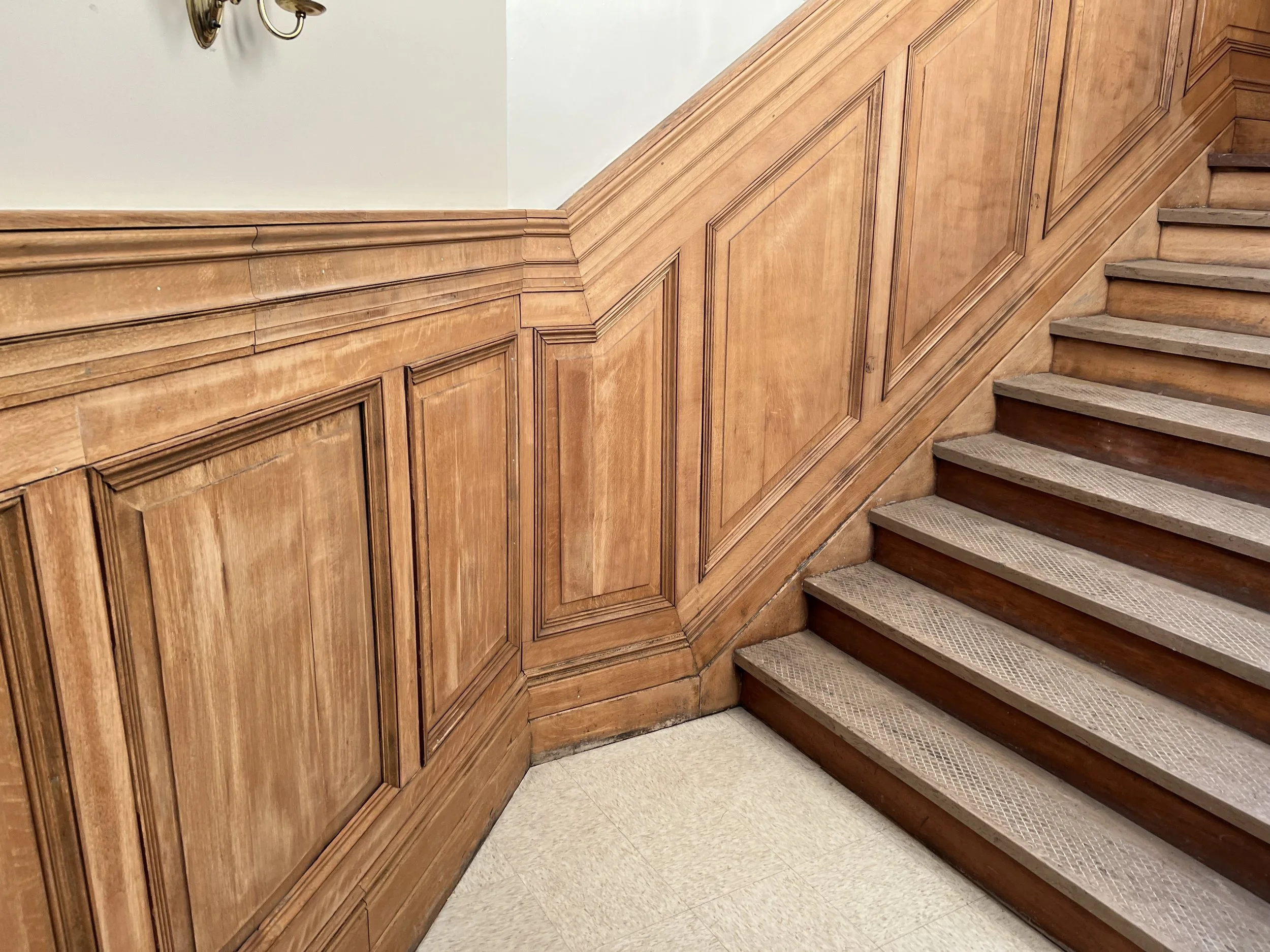 Wood-paneled staircase after laser stripping with vinyl stair treads, wooden wainscoting, and a wall-mounted light fixture.