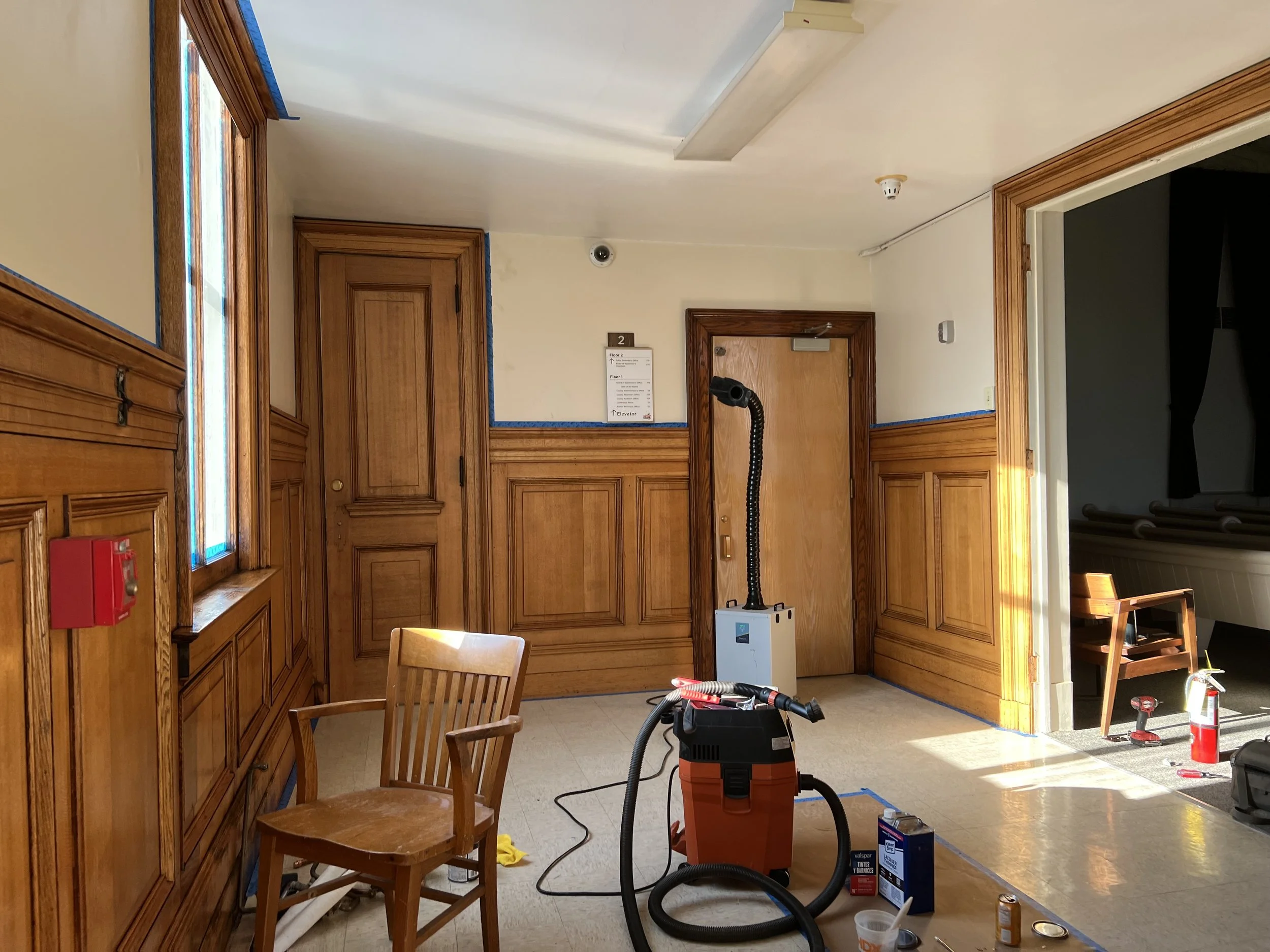 A room undergoing renovation or cleaning with wood-paneled walls, a chair, construction tools, and blue painter's tape along the ceiling edges.