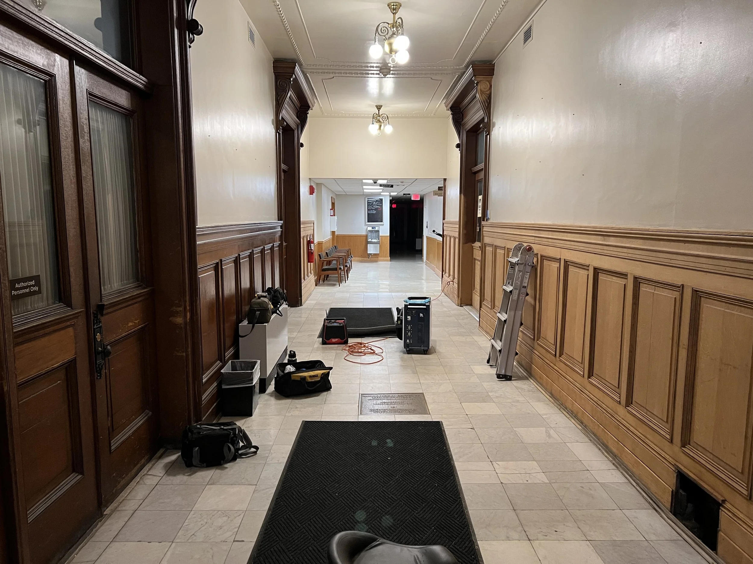Interior of a hallway with wood paneling and ornate crown molding, with equipment and bags placed on the floor, and ladders leaning against the wall.