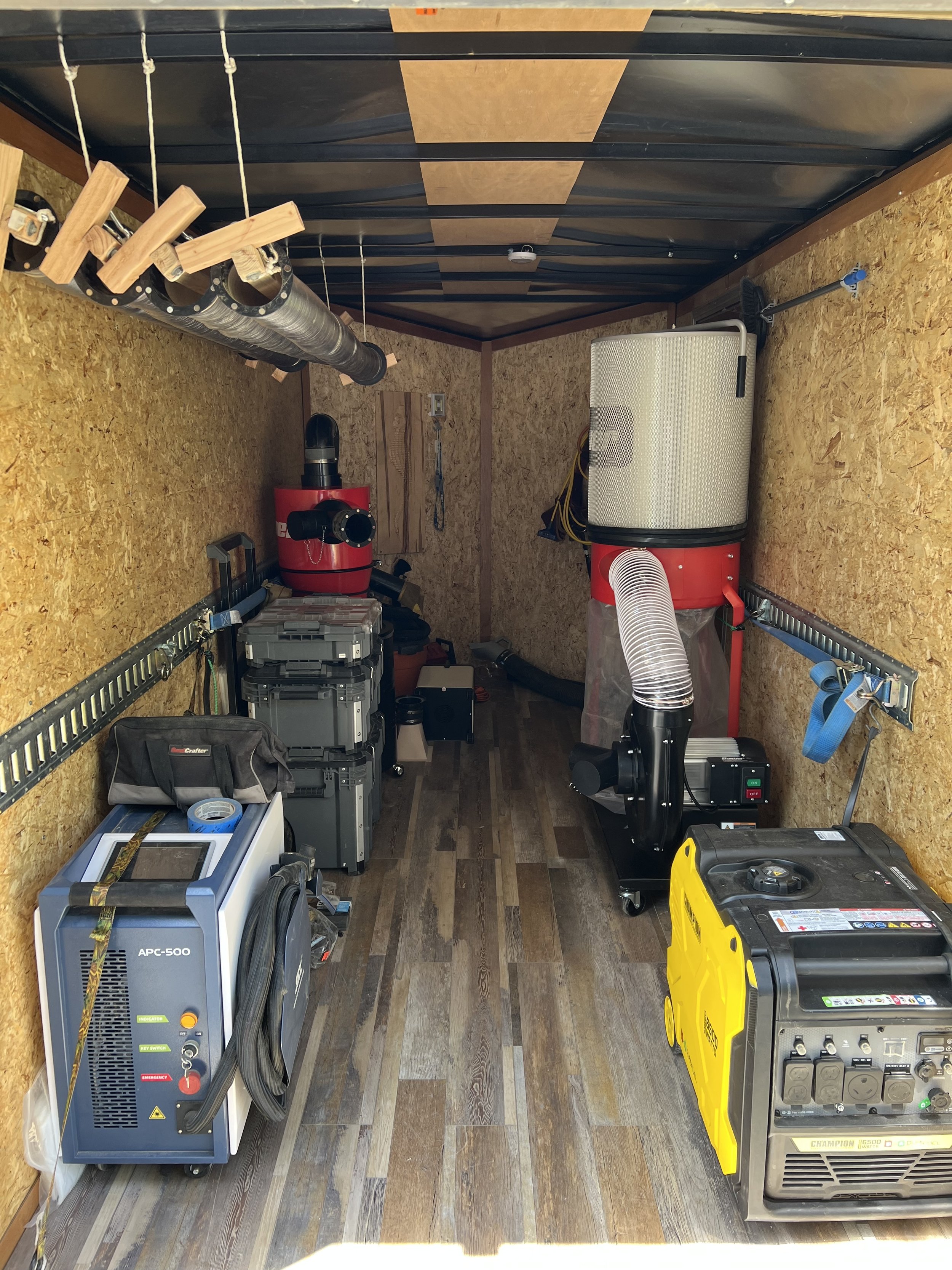 A small storage enclosed trailer with a wooden floor and plywood walls, containing various tools, equipment, and machinery including a dust collector, a generator, a vacuum, and storage containers.