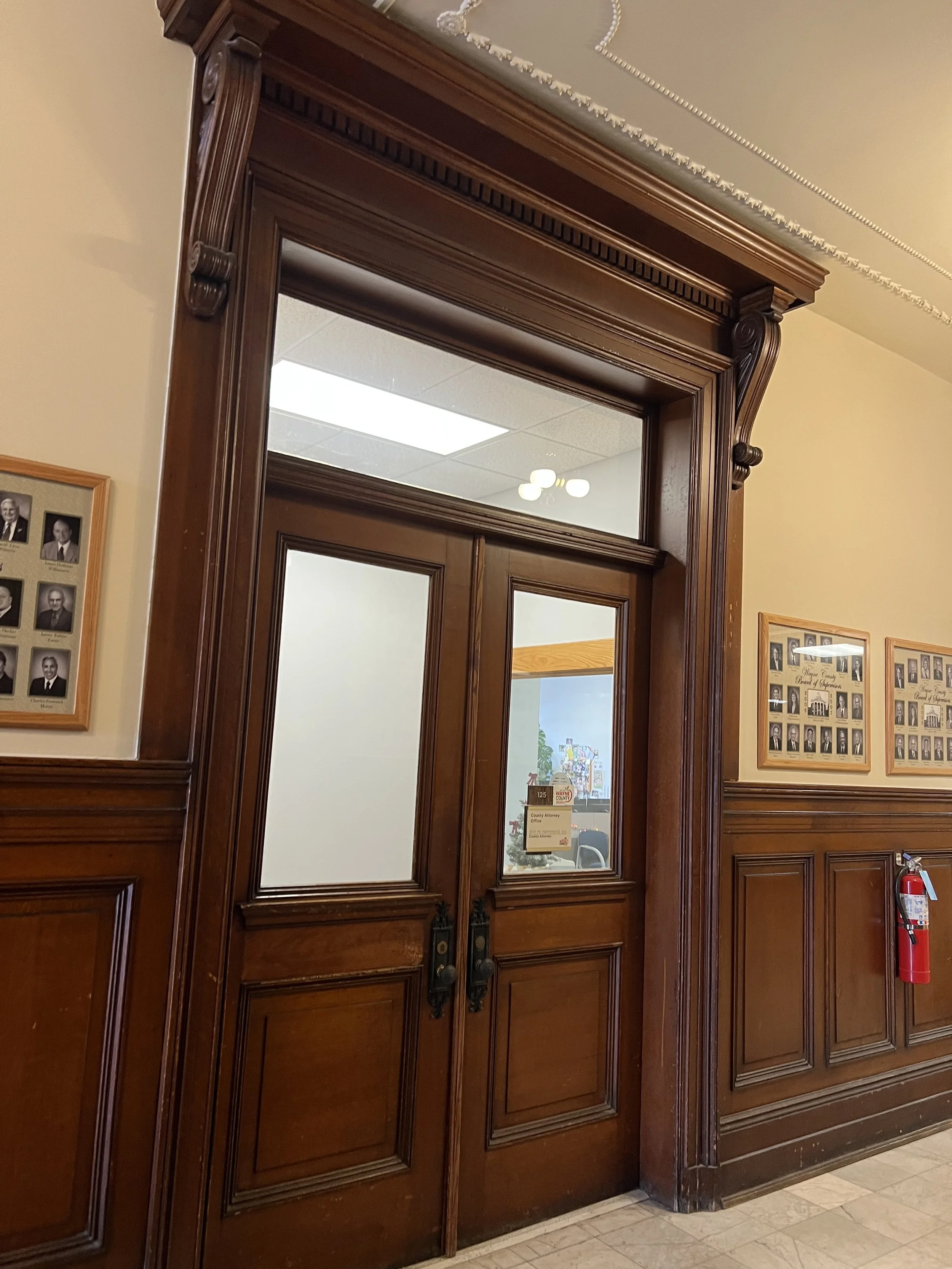 Wooden double doors with frosted glass panels and ornate trim, leading into an office or official building. A wall with framed photographs and a fire extinguisher are visible nearby.