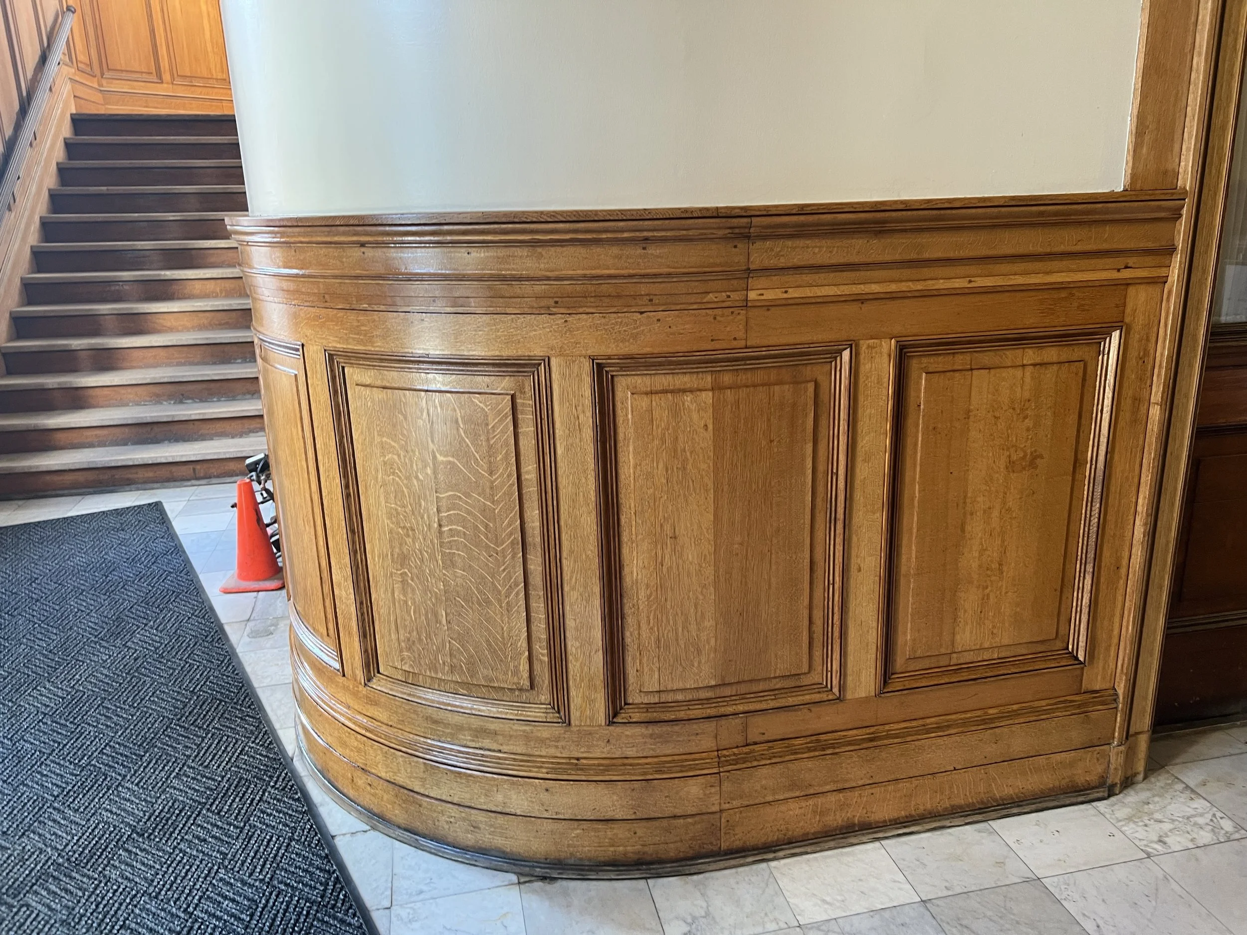 A curved wooden reception desk with detailed paneling in a building lobby. There are stairs and a carpeted floor area nearby, along with a small orange traffic cone.