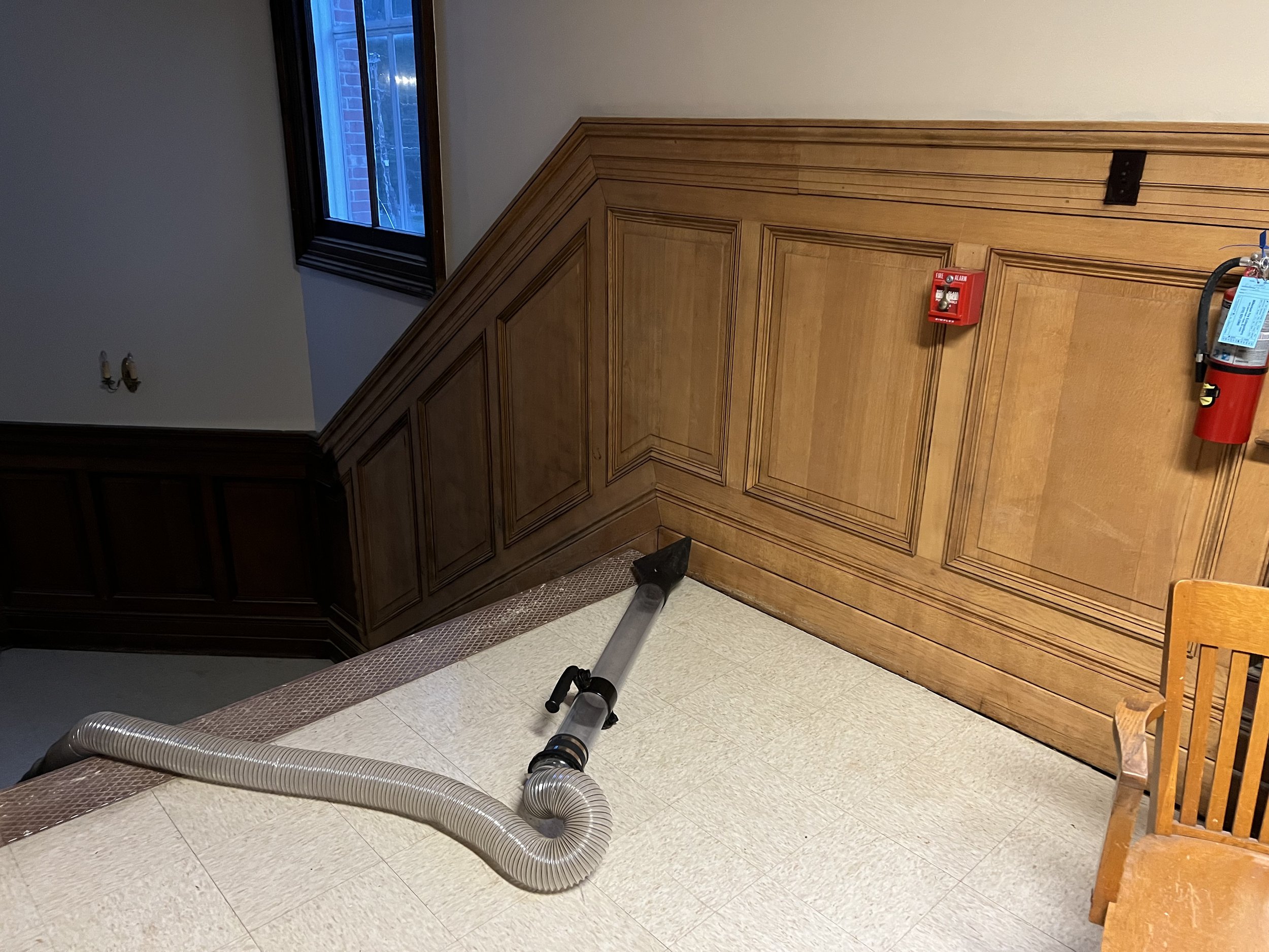 A vacuum hose on the floor in front of a wood-paneled wall with a fire alarm and fire extinguisher. There is a window with a blue view and a wooden chair to the right.