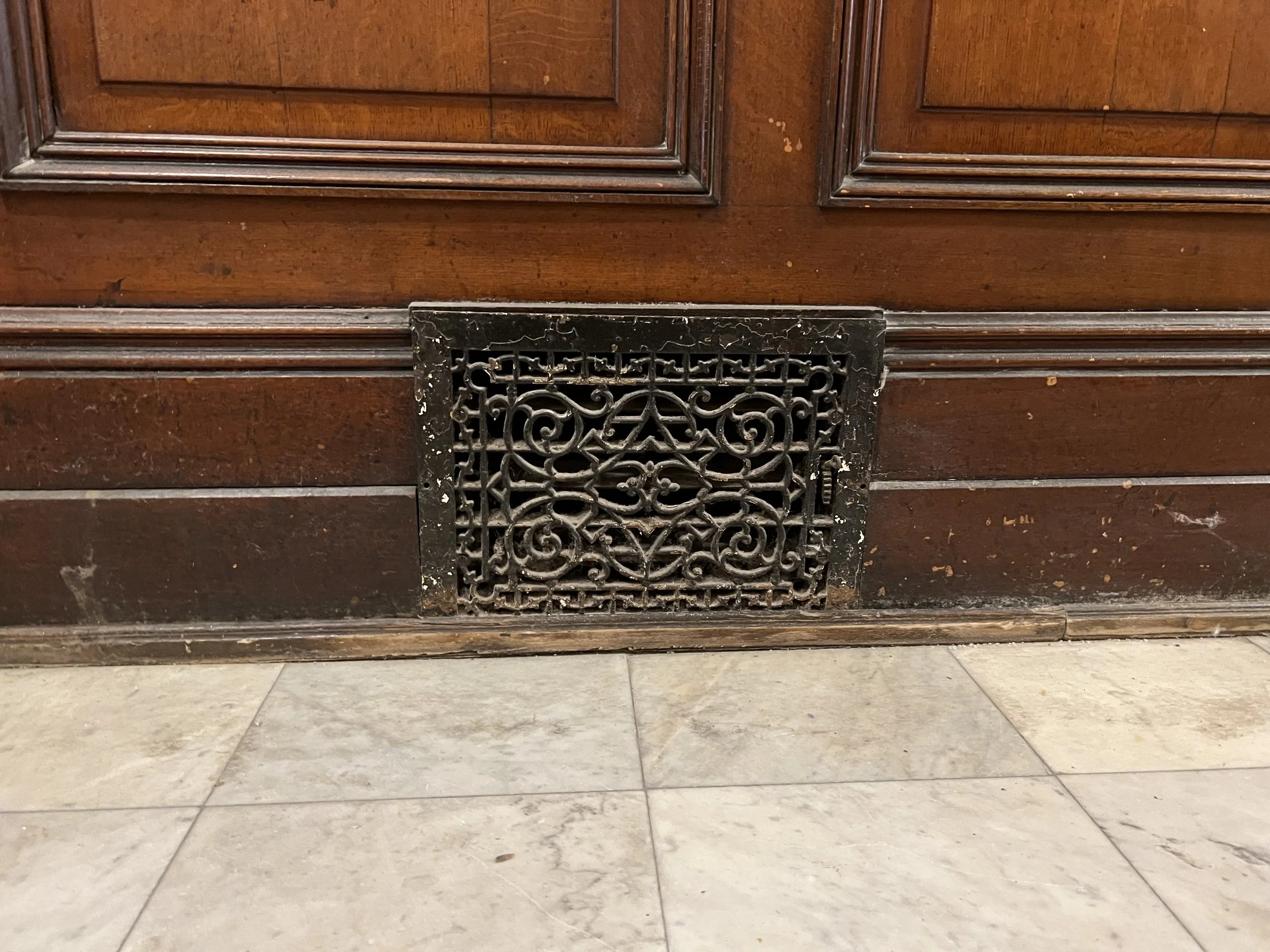Close-up of antique wooden furniture with black decorative metal vent at the bottom, on a gray tiled floor.