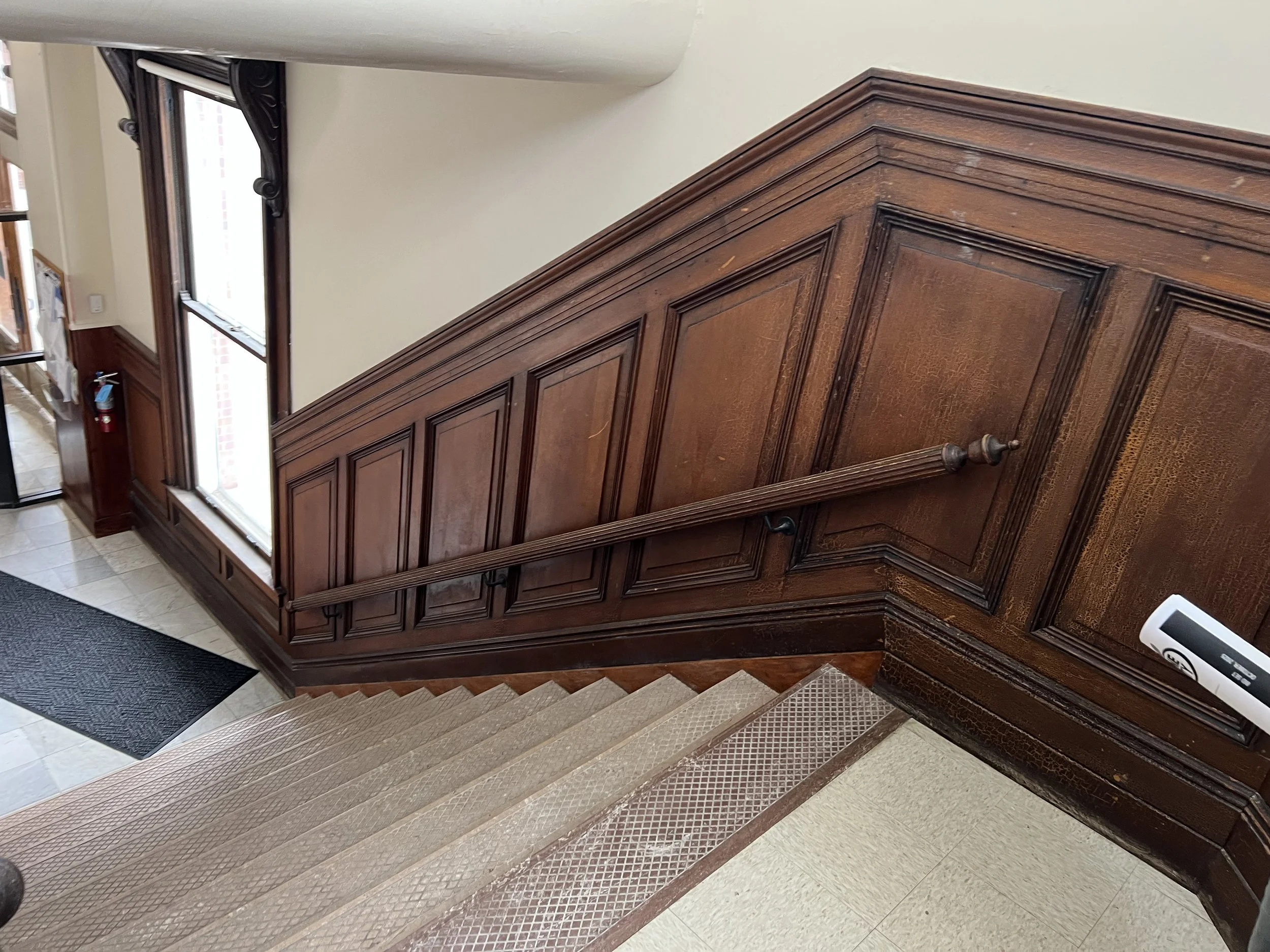 Inside a building with a staircase featuring wooden paneling on the wall and a handrail. There are large windows with decorative trim, a fire extinguisher on the wall, and a black rubber mat at the staircase entrance.