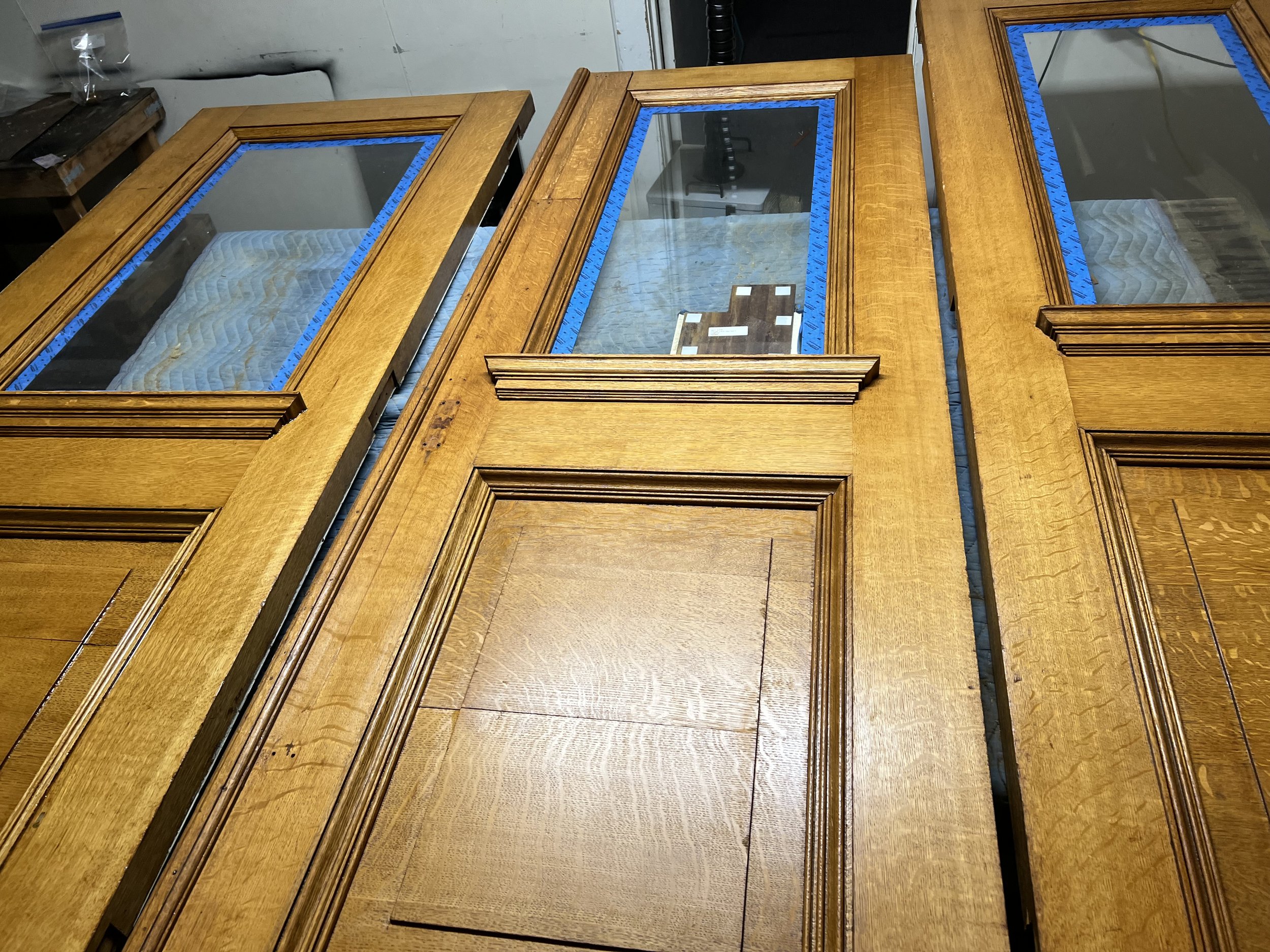 Three wooden dressers with glass tops, blue painter's tape along the edges, and a mattress underneath, in a workshop or storage room.