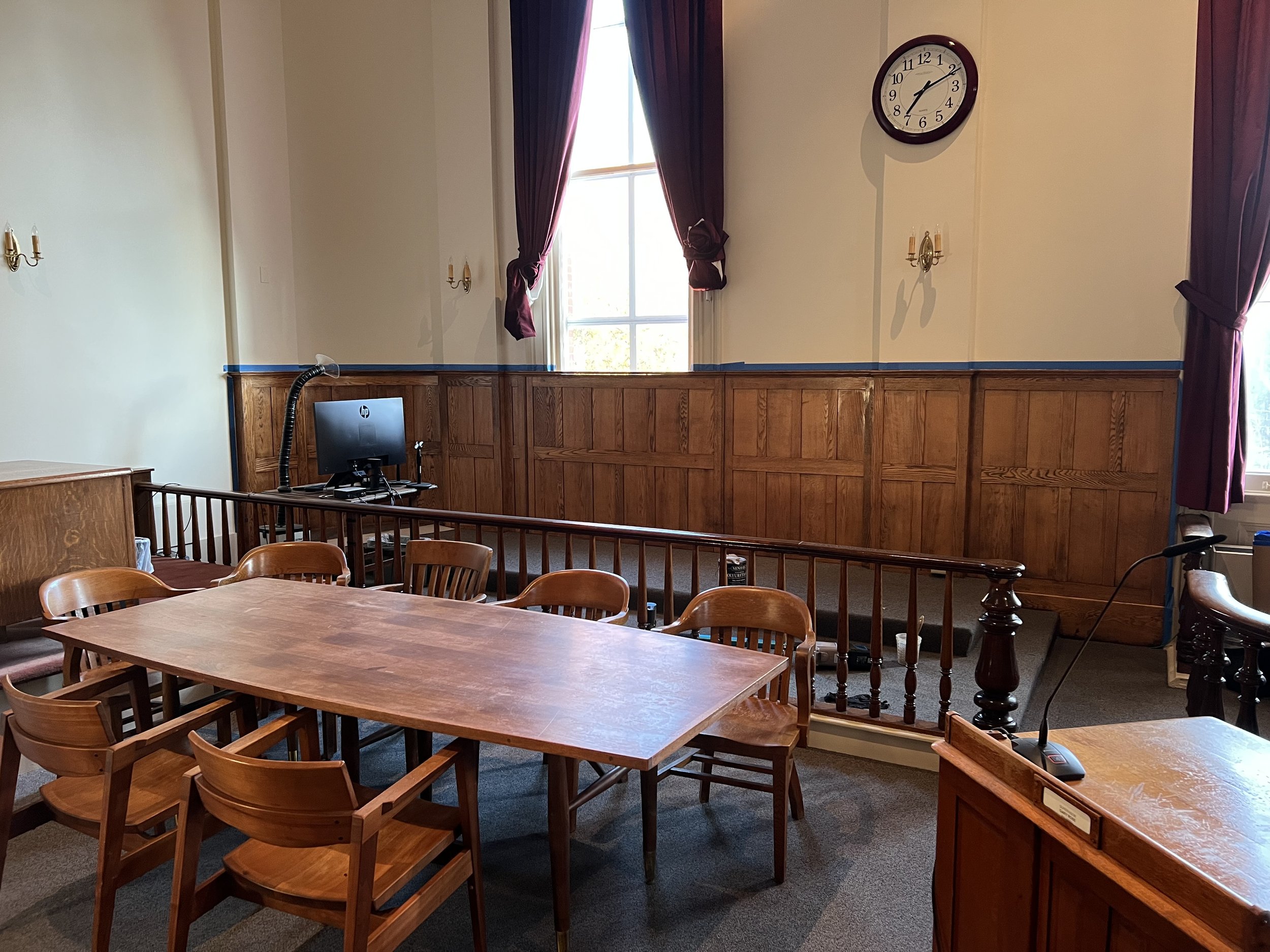 A room with wooden paneling on the lower walls, large window with purple curtains, and a wall clock showing 2:11. Wooden tables and chairs are arranged in the room, with a computer monitor and microphone on one table. The room appears to be a formal meeting or conference room.