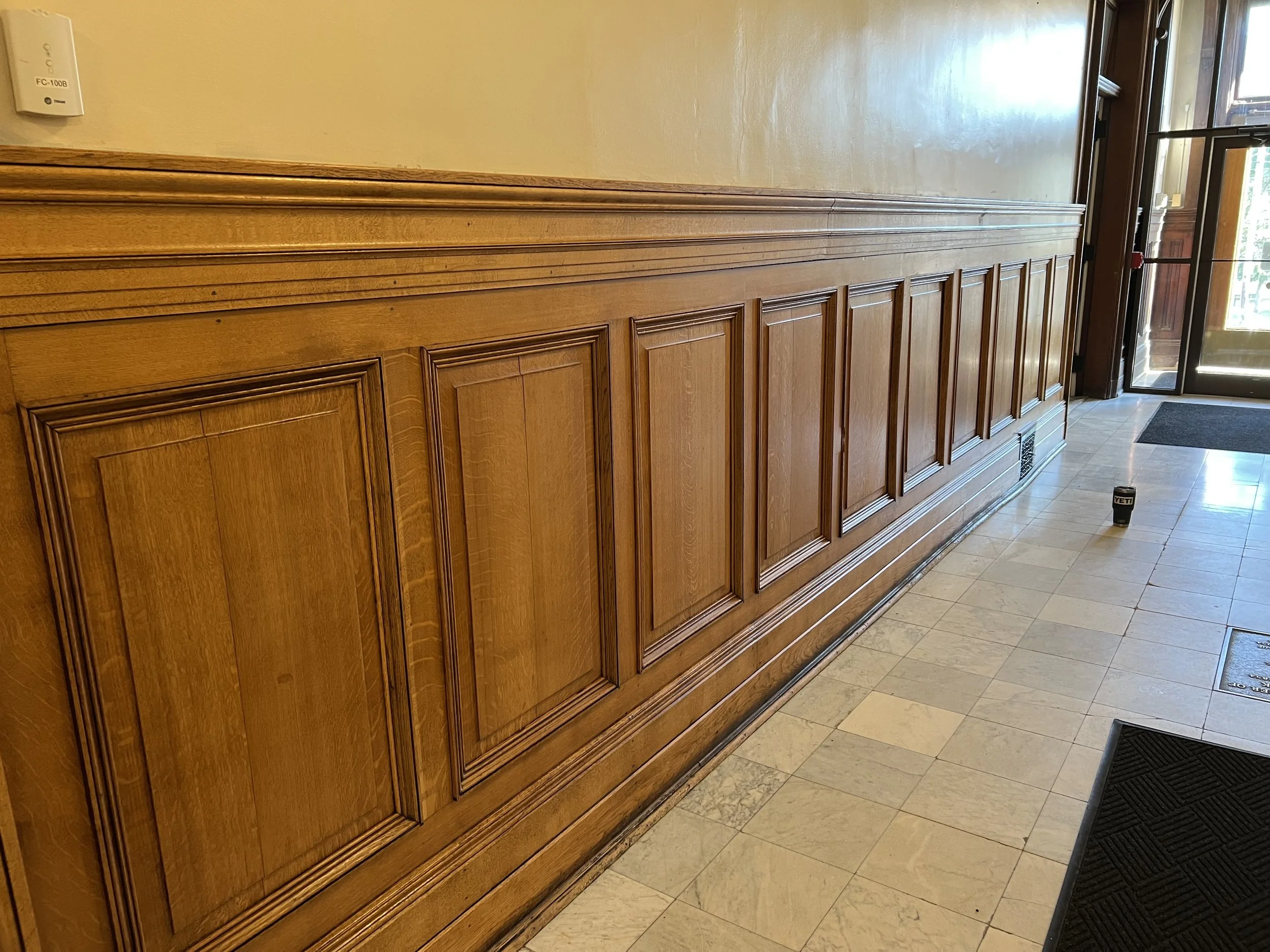 Wood-paneled wall inside a building, with a partially open glass door to the right showing the outside. There is a YETI tumbler on the floor near the wall.