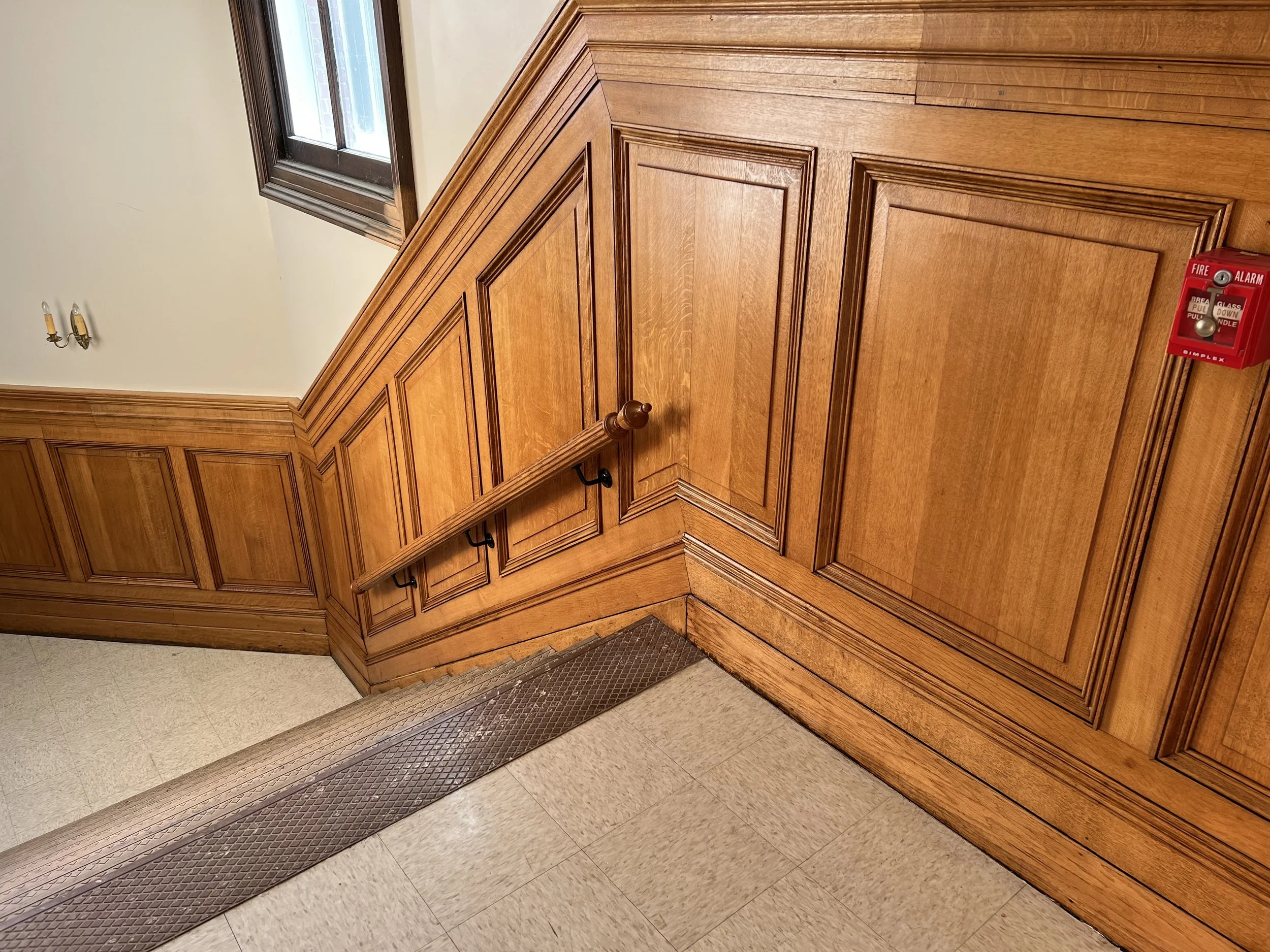 Wood-paneled elevator with beige walls, a window, emergency alarm, and a metal threshold at the bottom.