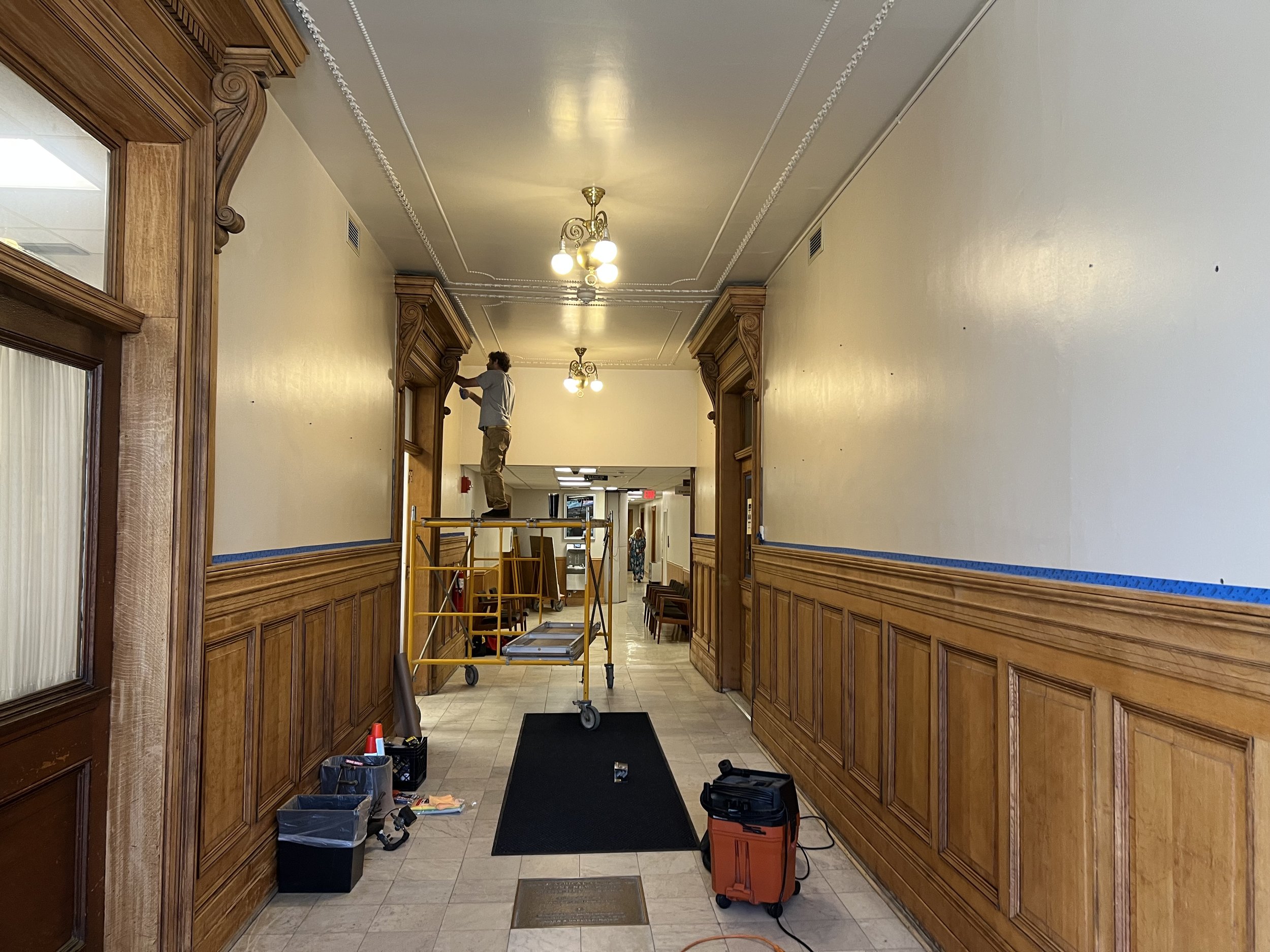 A person fixing or installing a light fixture on the ceiling in a hallway with wooden wall paneling, using a ladder and surrounded by tools and boxes.