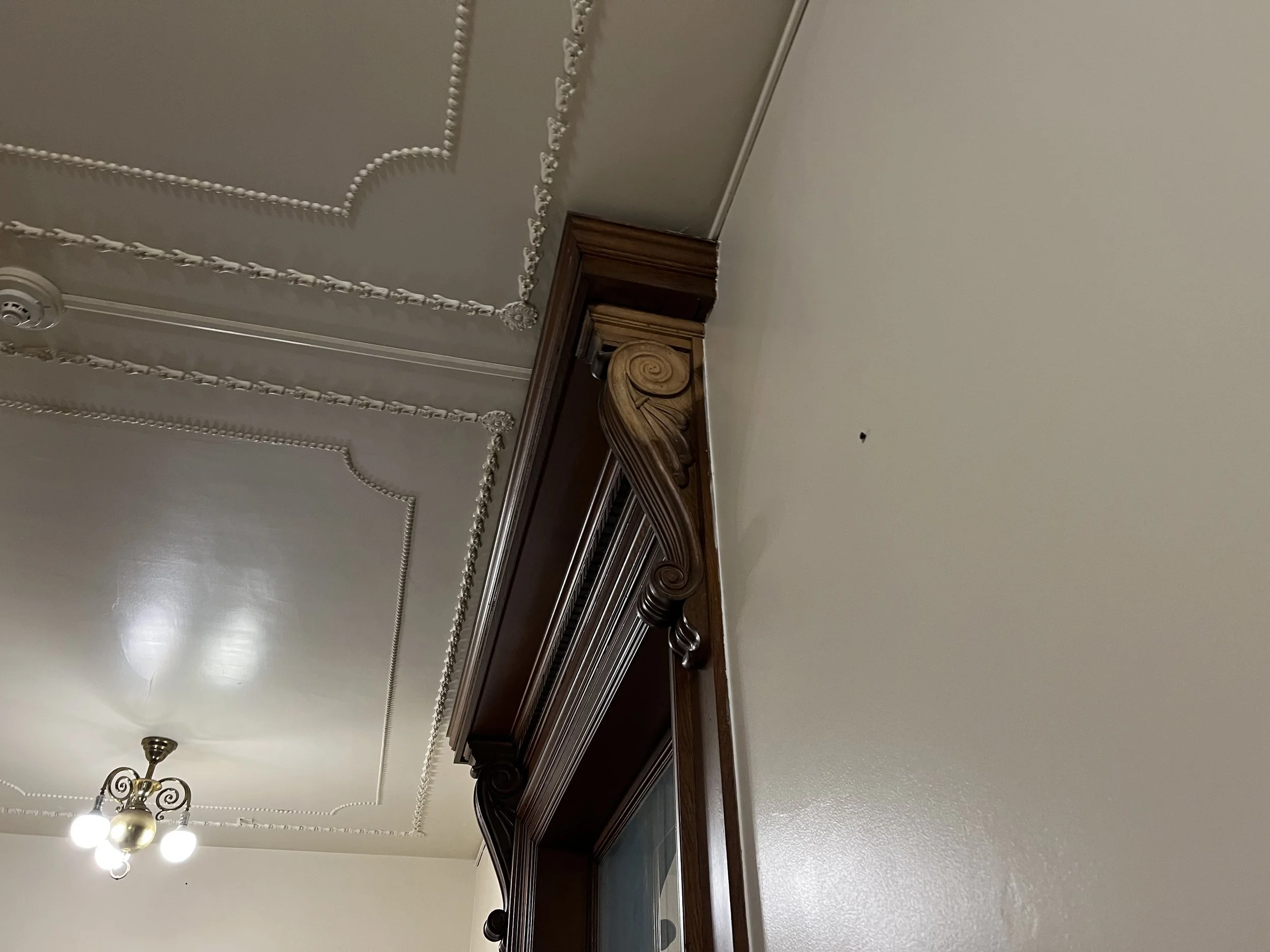 The image shows a ceiling with decorative molding, a section of a fireplace mantel made of dark wood with intricate carvings, and two ceiling light fixtures with multiple bulbs and ornamental details.