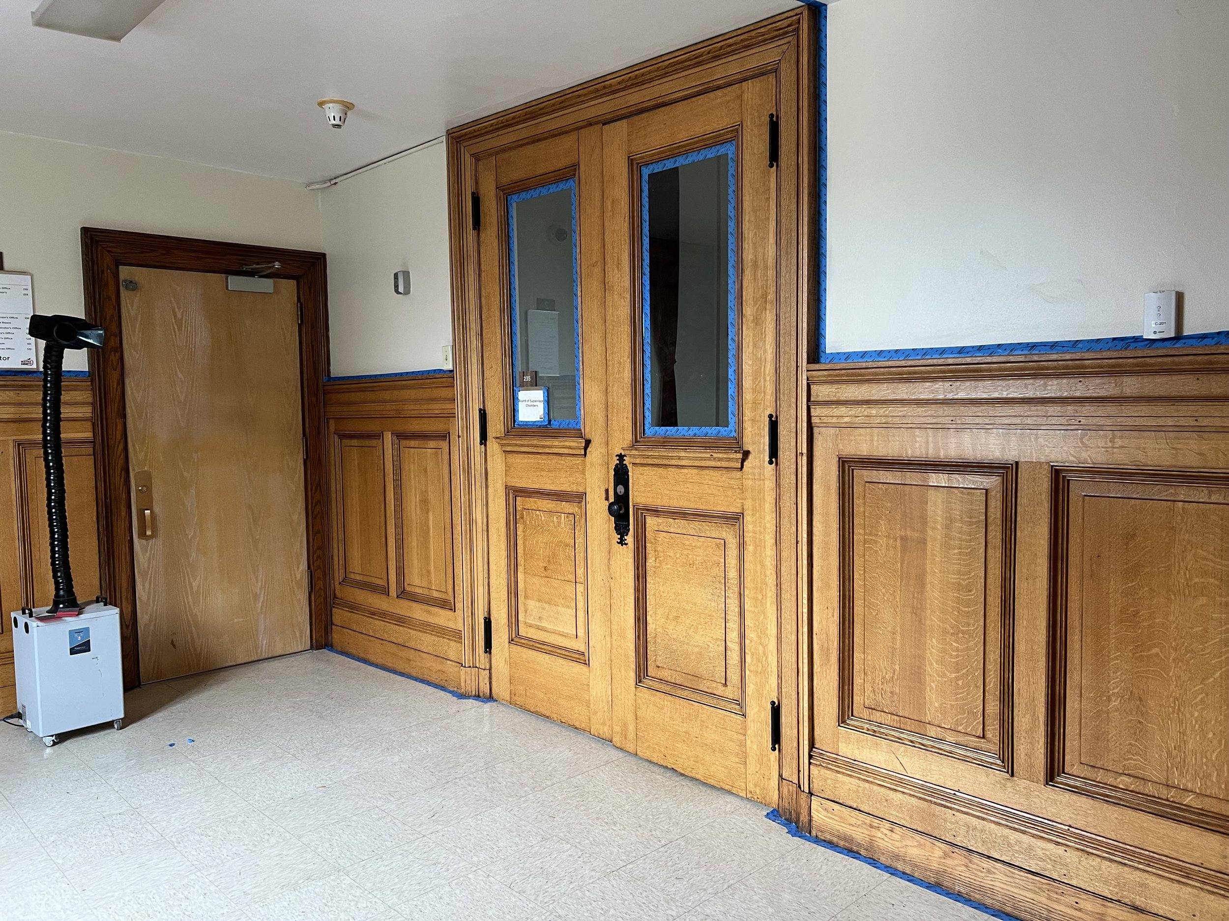 Interior of a room with wood-paneled walls and a closed wooden door. Blue painter's tape outlines and frames around a set of glass cabinet doors and parts of the walls. A smoke detector on the ceiling and an air purifier on the floor are visible.