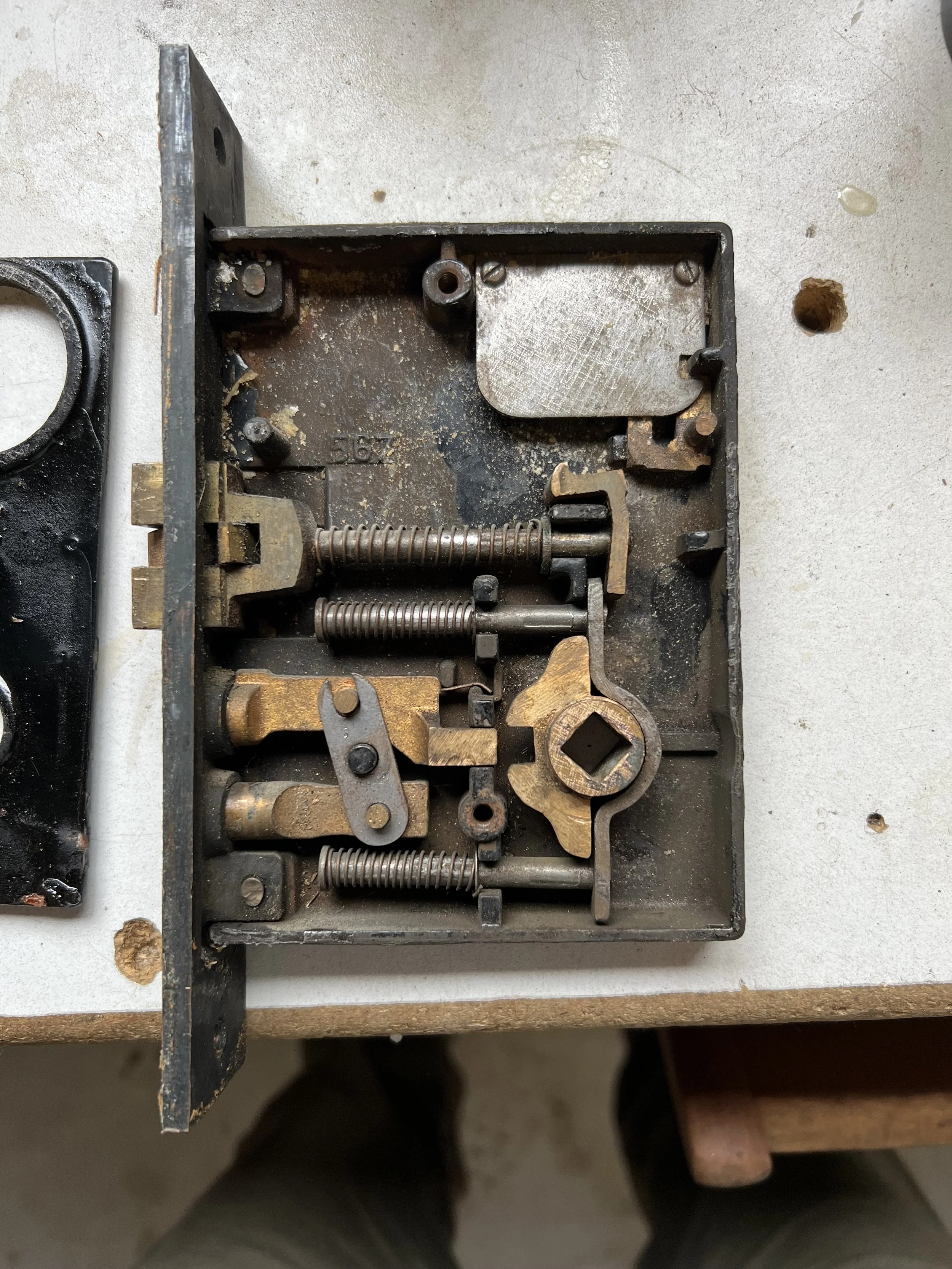 A disassembled vintage mechanical lock mechanism with springs, levers, and a square key opening inside a black rectangular metal casing.