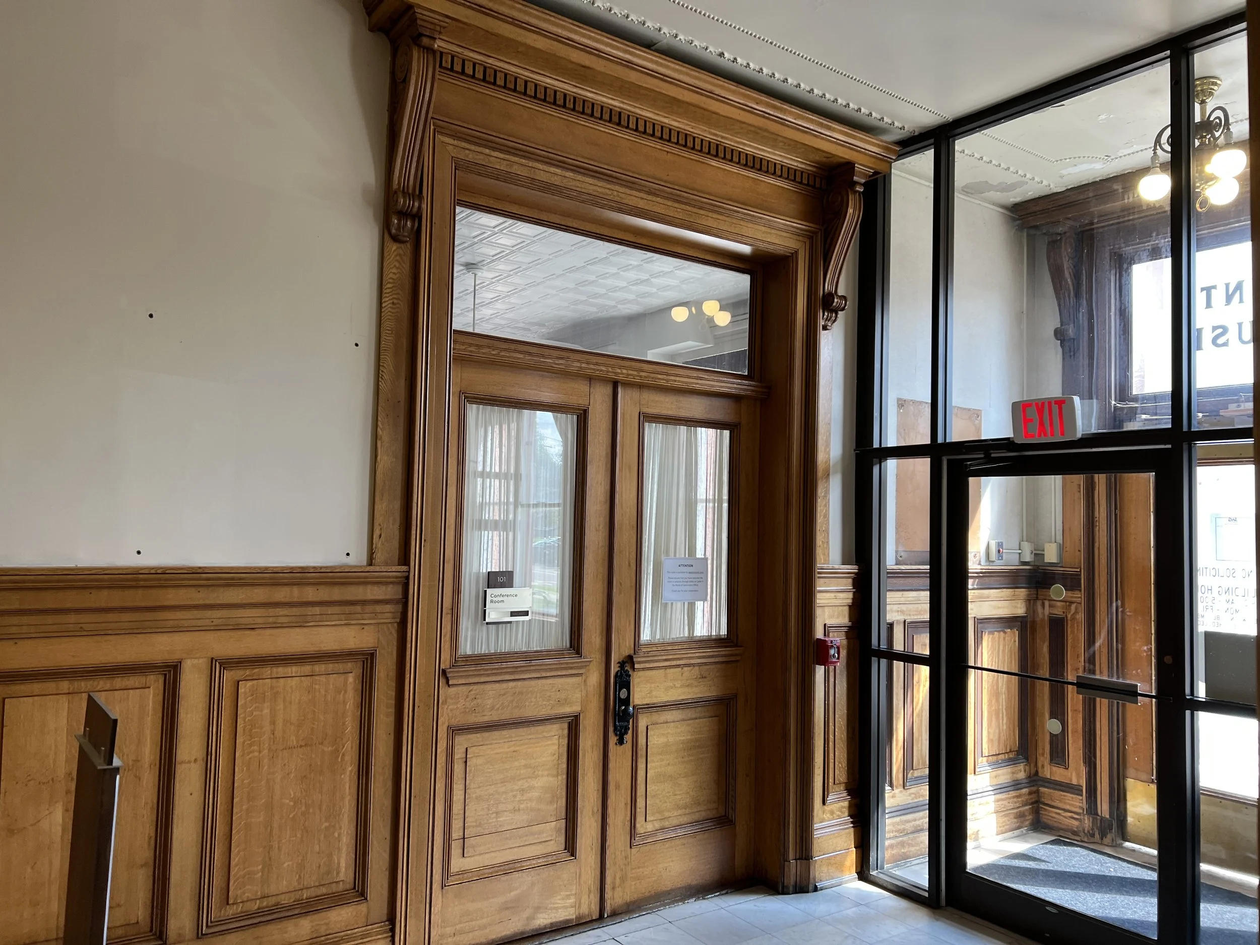 Wooden double doors with glass panels, an adjacent glass entrance door with an 'EXIT' sign above, and a glass window showing a chandelier and ceiling details inside the building.