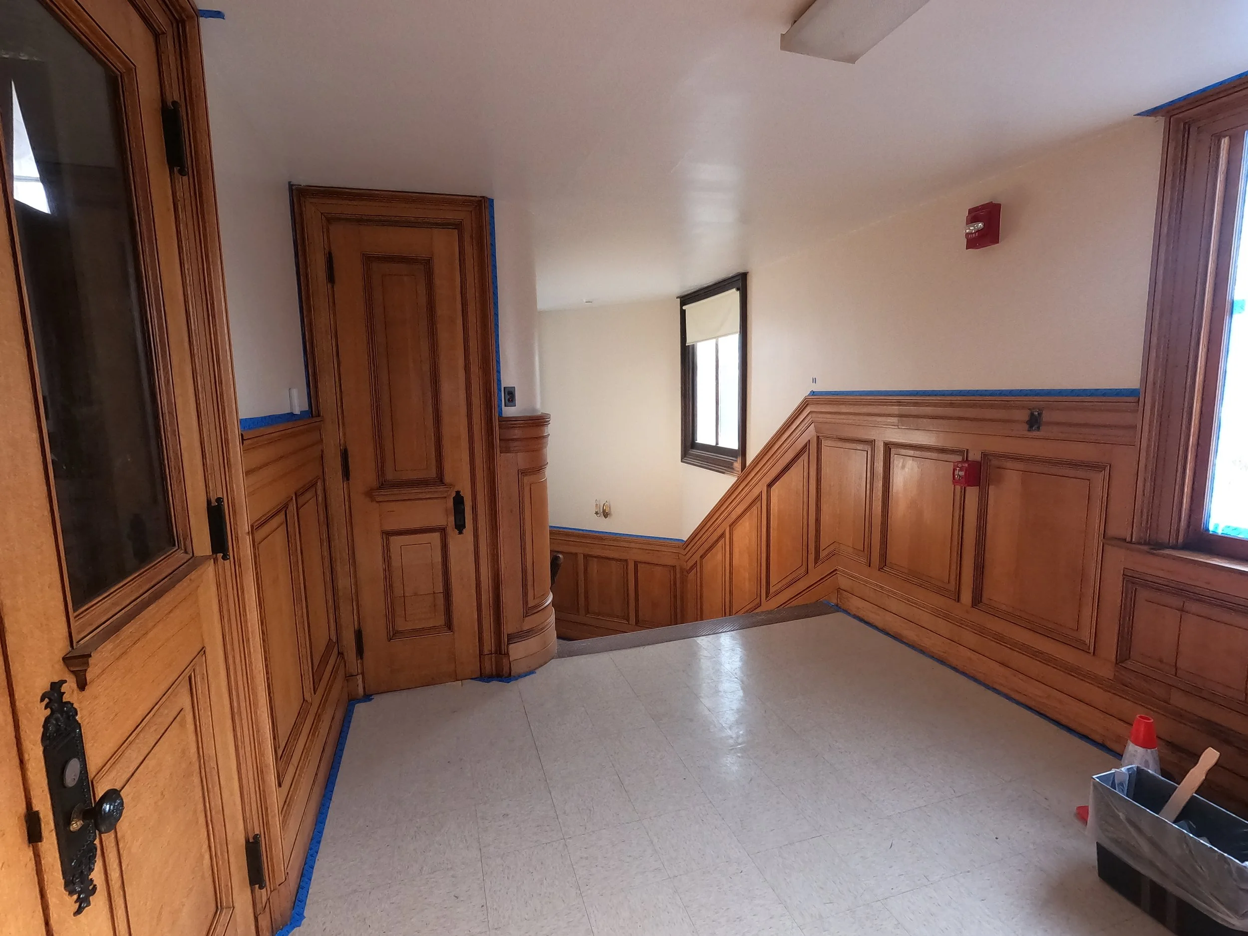 Interior of a room with wood-paneled walls and cabinets, a window with a shade, a fire alarm on the wall, and construction supplies on the floor, indicating ongoing renovation.