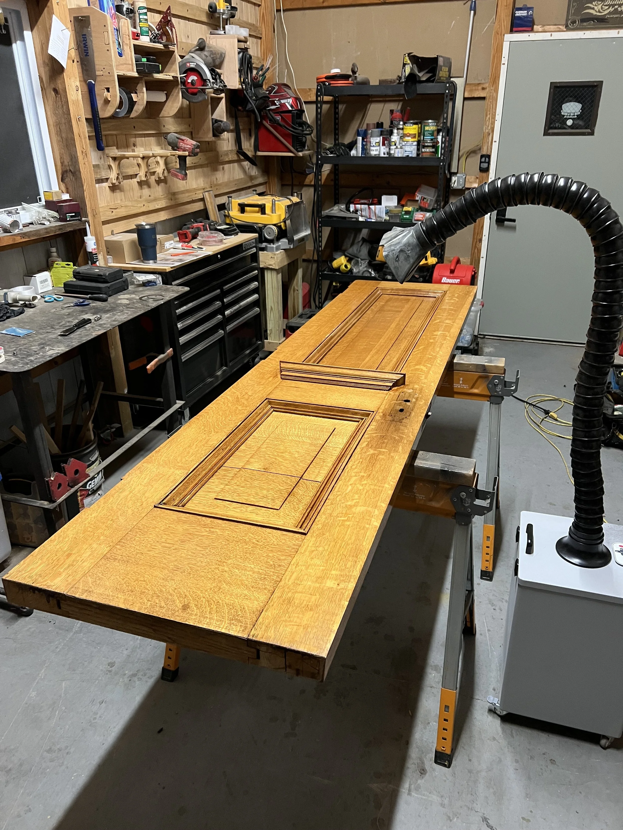 A woodworking workshop with a large wooden table, various tools and supplies, a workbench, and shelves with paint cans and hardware. A fume extractor extends over the table.