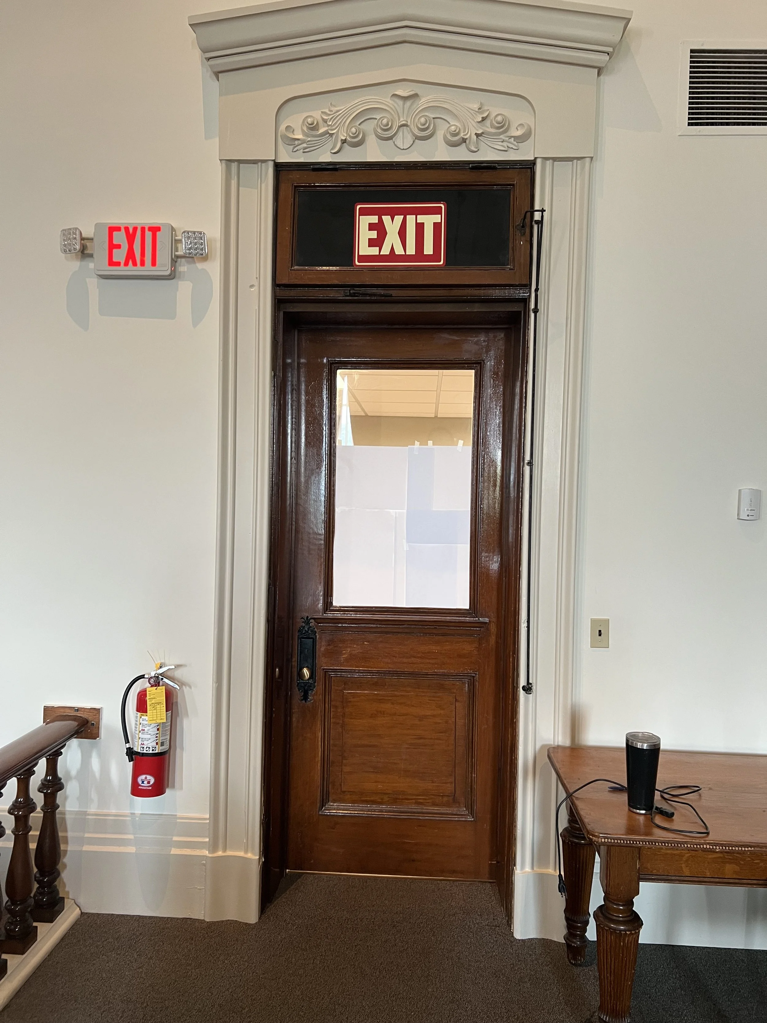 Wooden door with glass window at the top, decorated with ornate white molding above, inside a room. Two red exit signs are above and to the left of the door. A fire extinguisher hangs on the wall to the left of the door, and a wooden table with a black device and cords is to the right. A small window vent is in the wall to the right of the door.