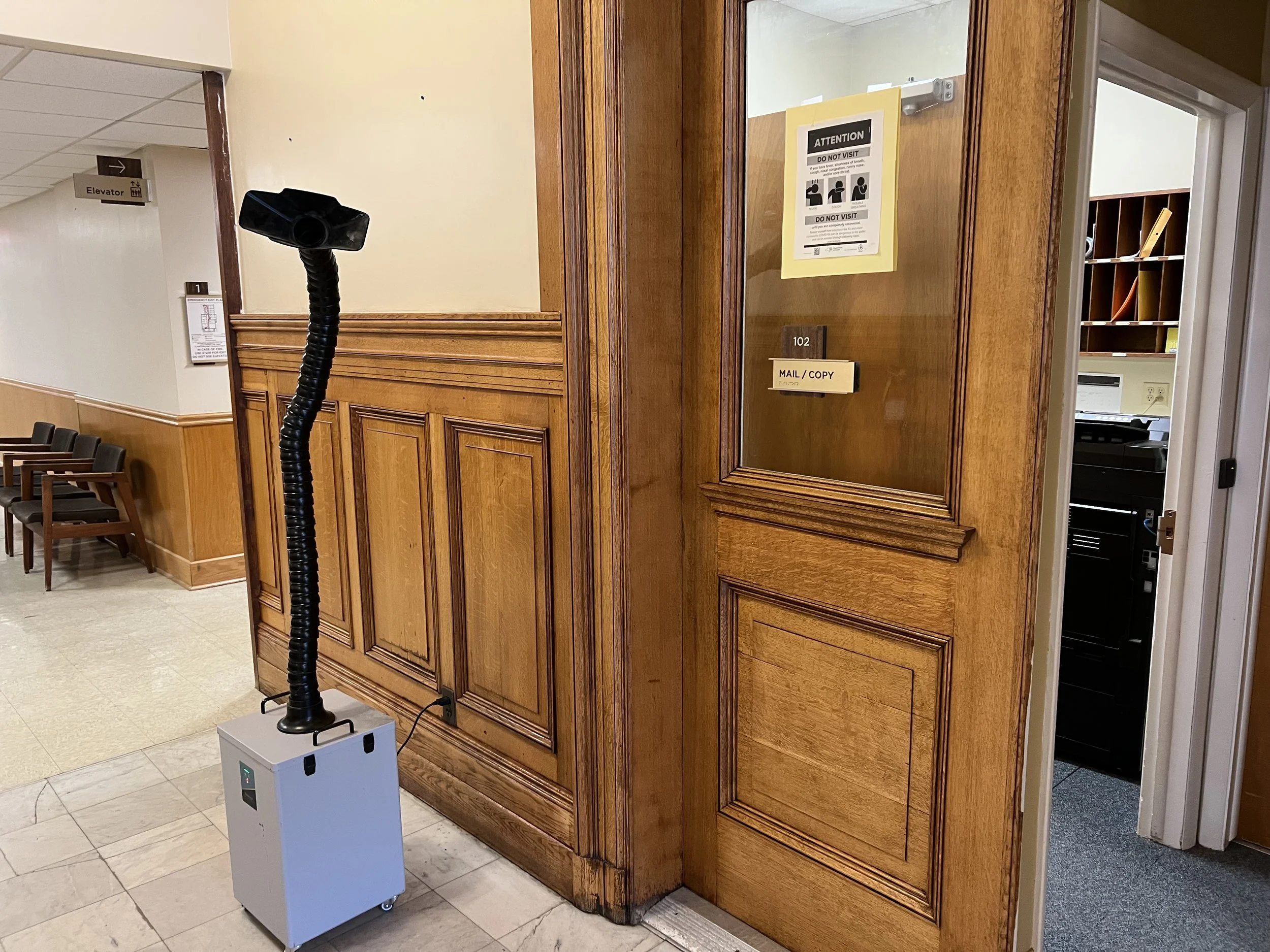 A portable air purifier with a flexible black tube hose attached, positioned in front of a wooden-paneled door marked with the number 102 and a mail/copy slot. Signs on the door warn against visiting. In the background, chairs are arranged along a wall near a corridor with an elevator sign.