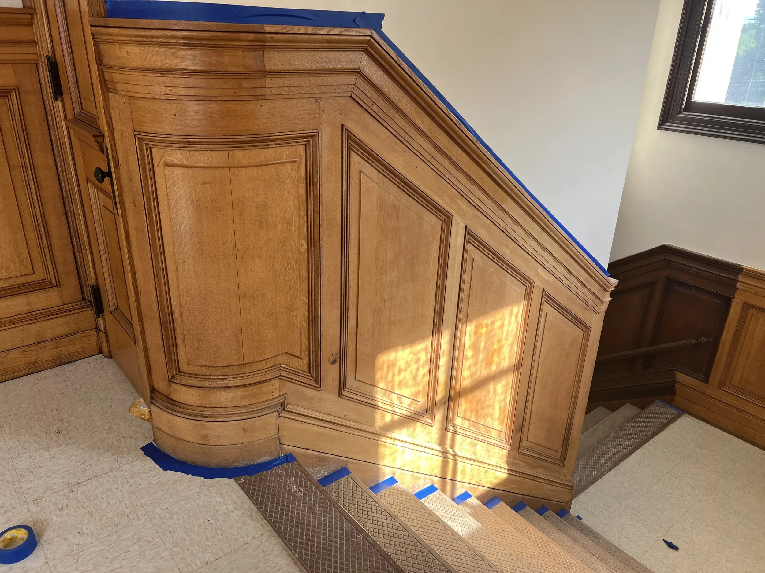 Close-up view of wooden custom cabinetry with detailed molding, in a room with a large window and a staircase. The cabinetry is partially taped, indicating ongoing renovation or finishing work.