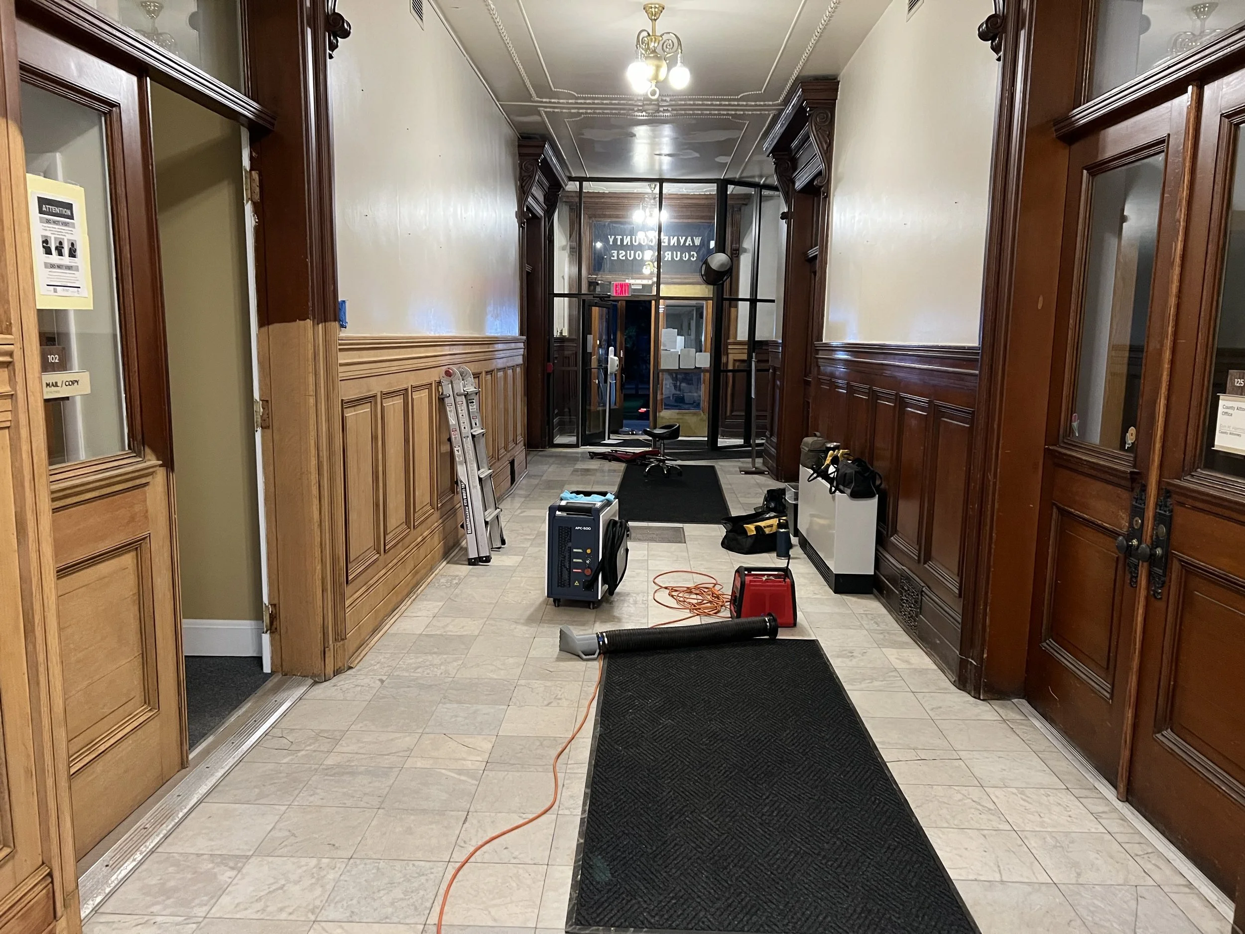 Empty hallway with wooden paneling, construction tools, a ladder, and a black rubber mat on beige tiled floor. Glass doors with a sign reading 'WAYNE COUNTY COURT HOUSE' are at the end of the hallway, with an exit sign above.