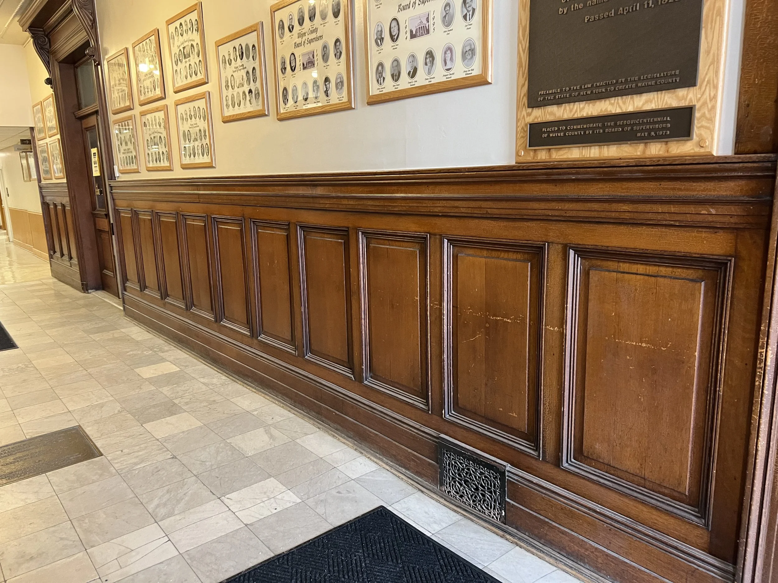 Interior of a government building with a wooden wainscoting and framed black-and-white photographs and plaques on the wall. The floor has beige tiles, and there are black vent covers near the base of the wall.