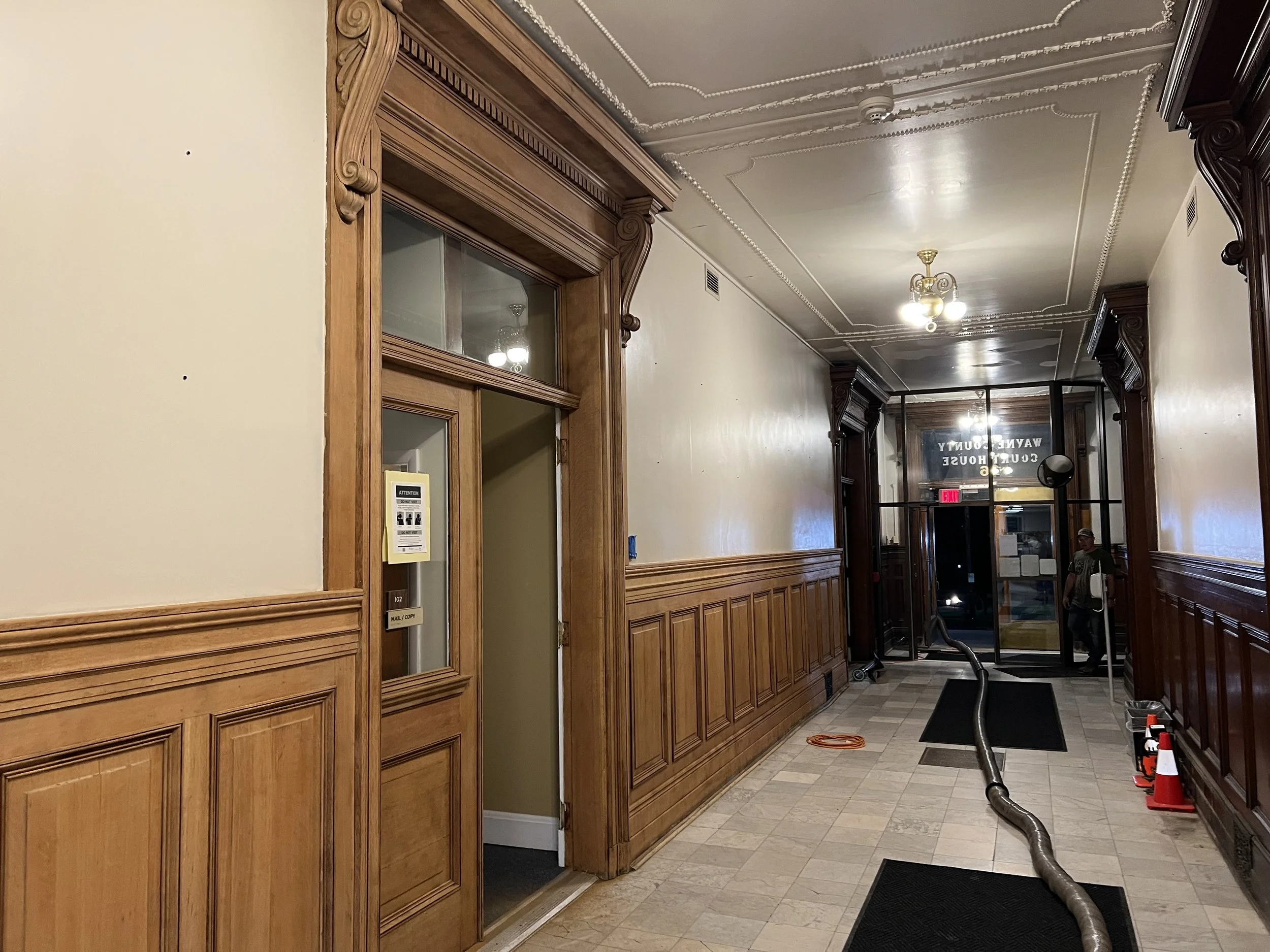 Interior hallway with wooden wainscoting and decorative trim, ceiling with ornate plaster molding, hanging light fixtures, and cleaning equipment such as a large vacuum hose, orange cones, and a water bottle.