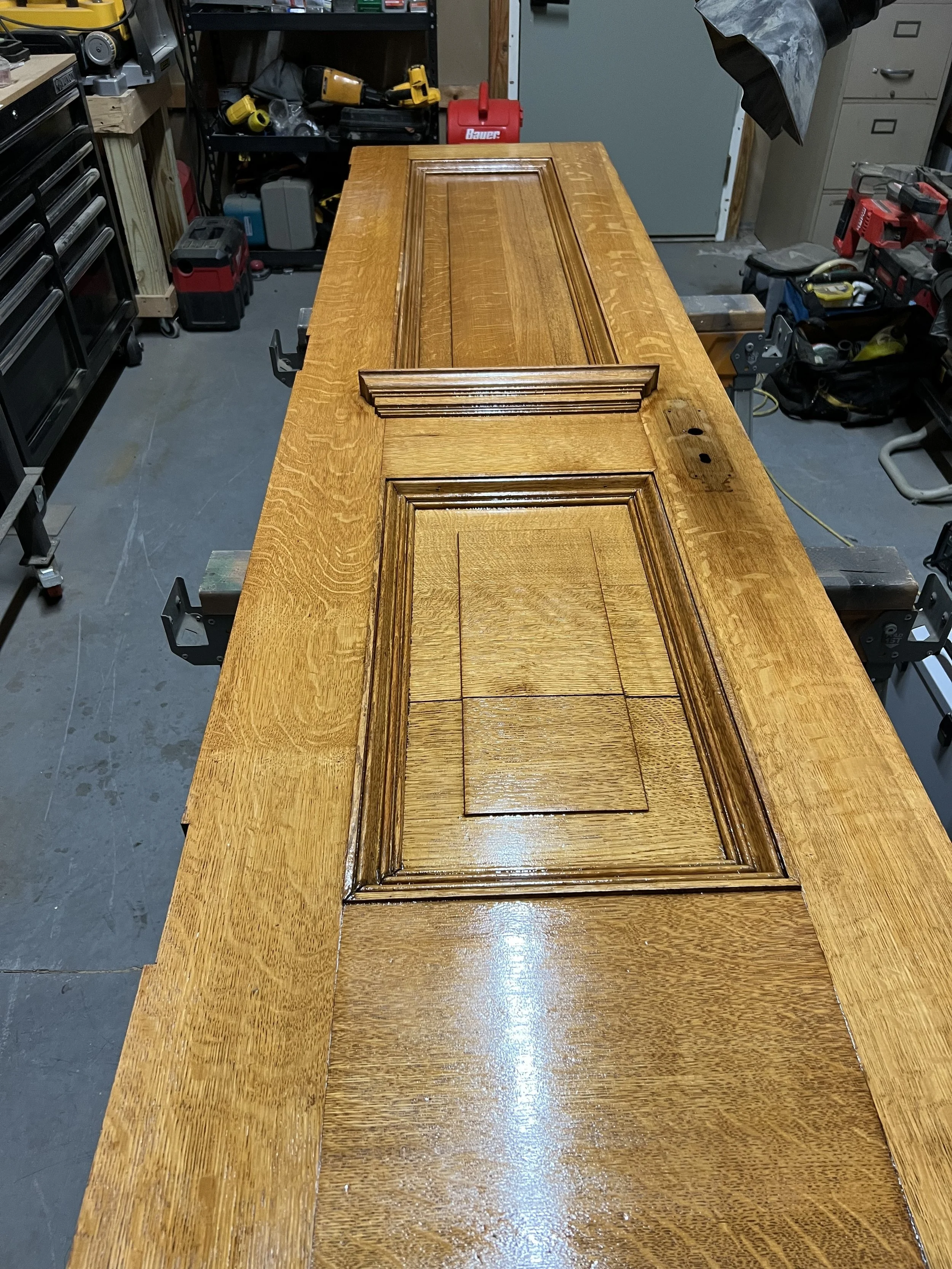 A wooden workshop table with a carved panel door resting on it, surrounded by various tools and workshop equipment.