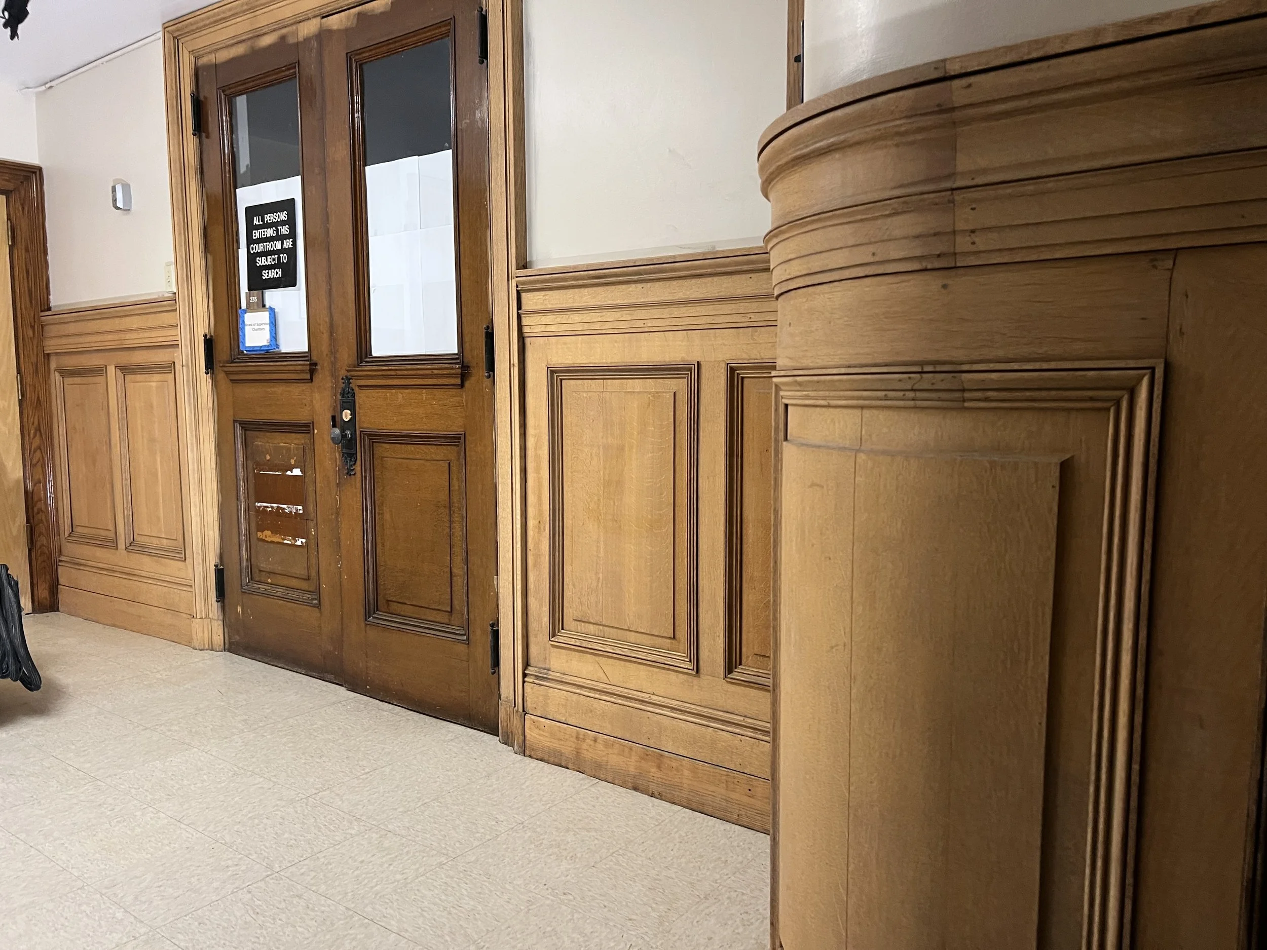 Wood-paneled courtroom with a set of doors and a small window. Sign on the door reads, 'All persons entering this courtroom are subject to search.'
