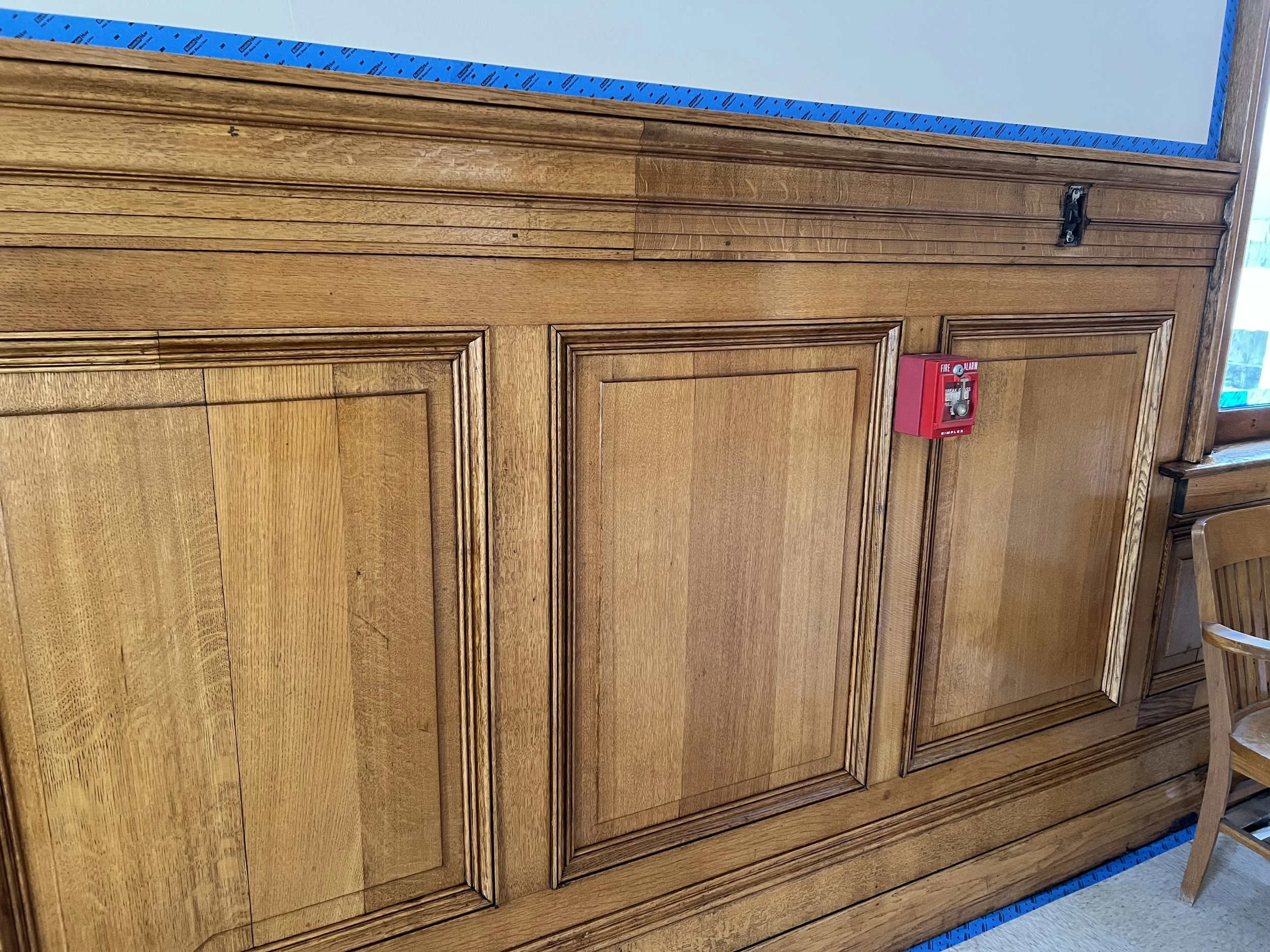 Wooden cabinet with a rectangular shape, panel doors, and crown molding, positioned against a wall with a blue painter's tape along its top edge, and a red fire alarm pull station mounted on its surface.