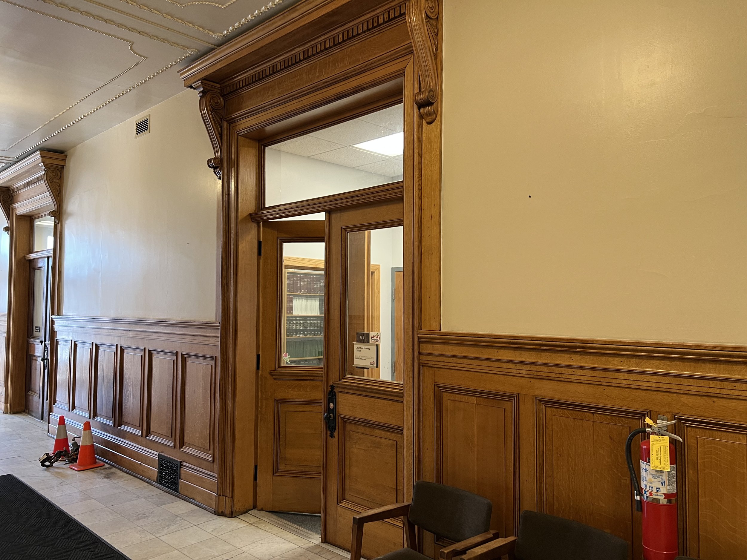 Wooden panel on the wall with two mirrors and a door, in a room with beige walls and tiled floor, with traffic cones and a fire extinguisher on the floor.