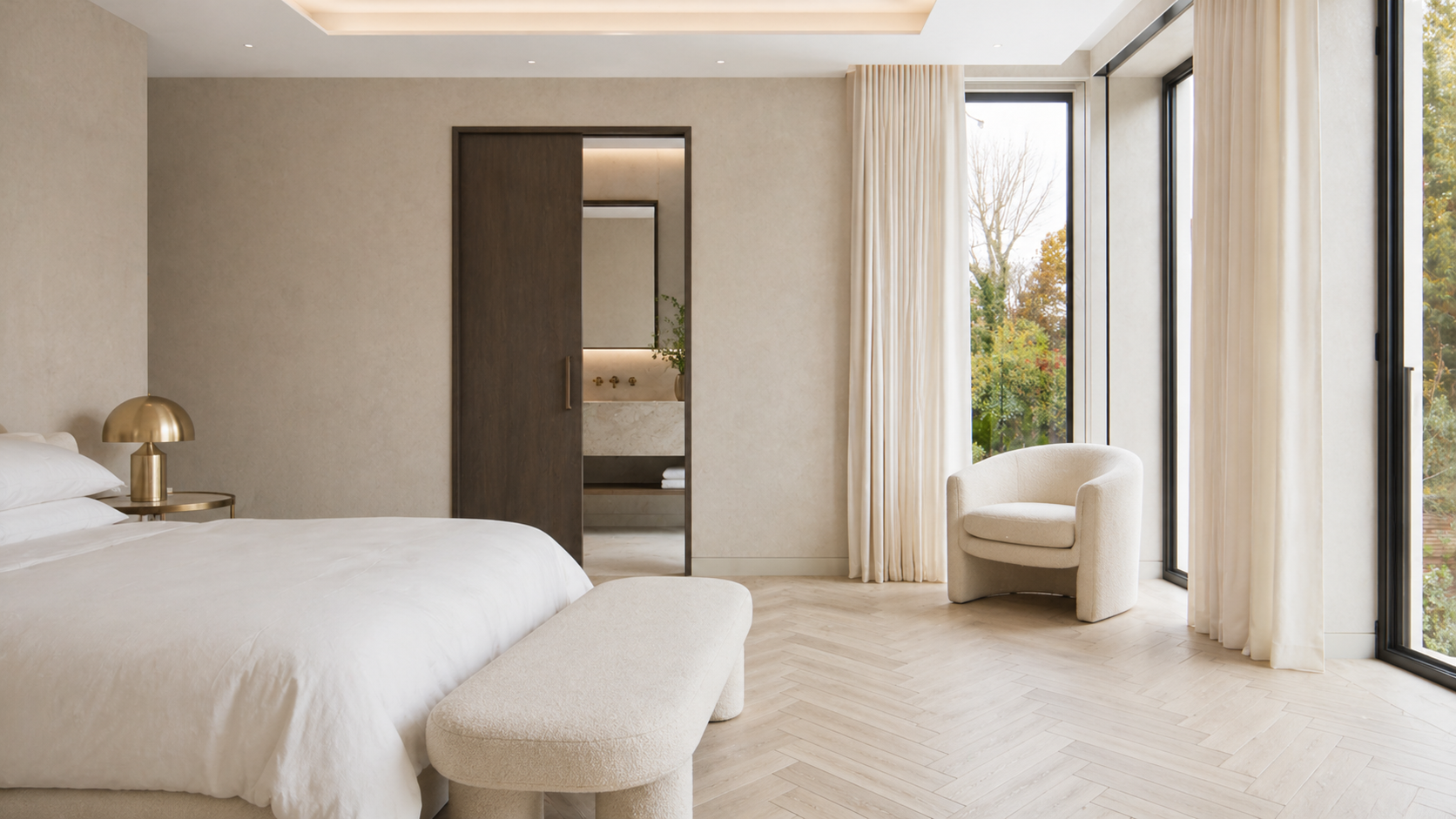 A modern bedroom with a large bed, a gold bedside lamp, a cream-colored armchair, and large windows with white curtains showing trees outside.