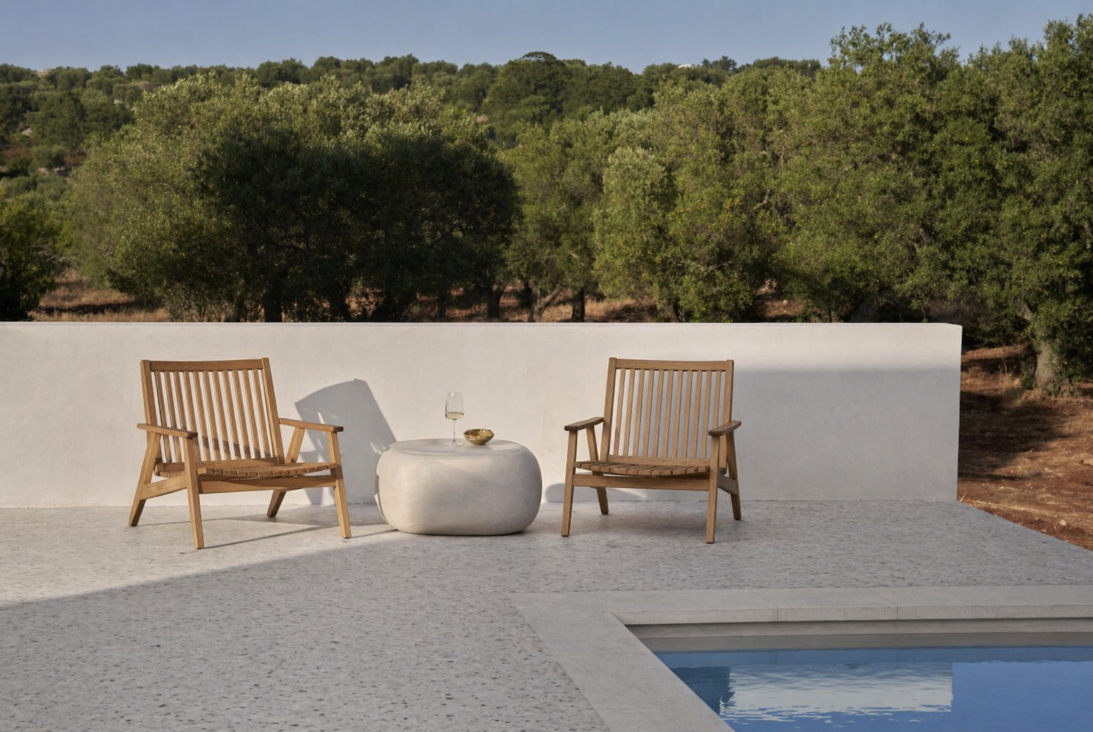 Two wooden chairs with armrests face each other on a concrete patio, with a round white table between them. On the table is a glass of white wine and a small bowl. In the background, there is a white wall and a landscape of trees and shrubs, with a small swimming pool in the foreground on the right.