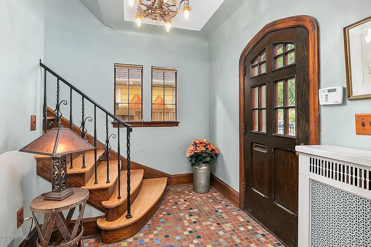 Entryway with a wooden front door, a small staircase with a black wrought iron railing, a colorful tiled floor, a vase with flowers, and a wall-mounted thermostat.