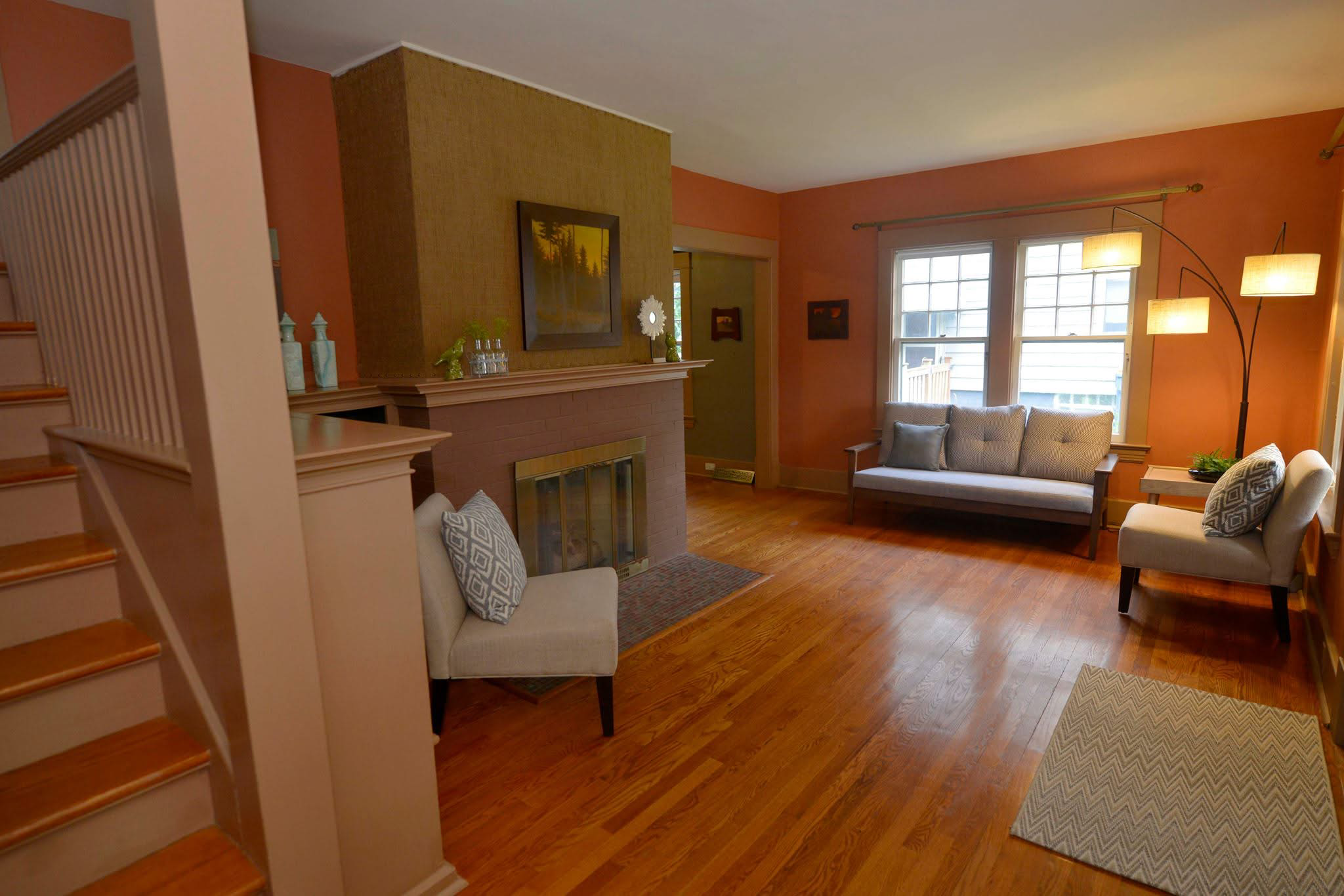 Living room with hardwood floors, beige and brown walls, a stone fireplace, beige sofas, and a large window with blinds.
