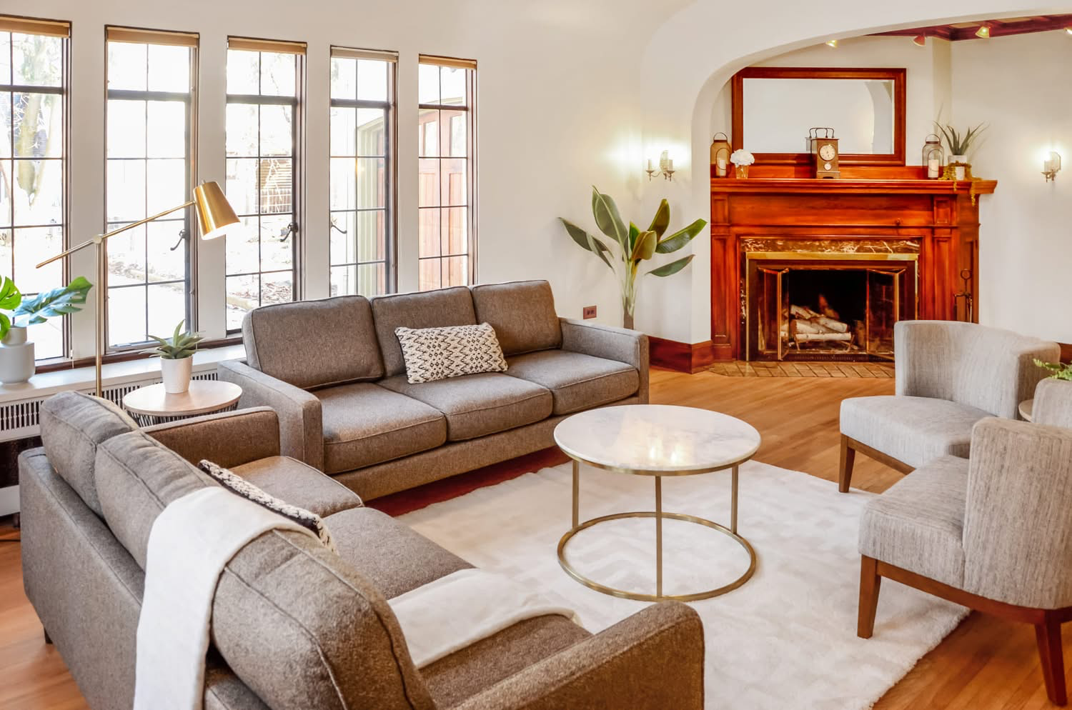 Living room with large windows, gray sofas, a white rug, a marble-topped coffee table, a fireplace with a wooden mantle, and indoor plants.