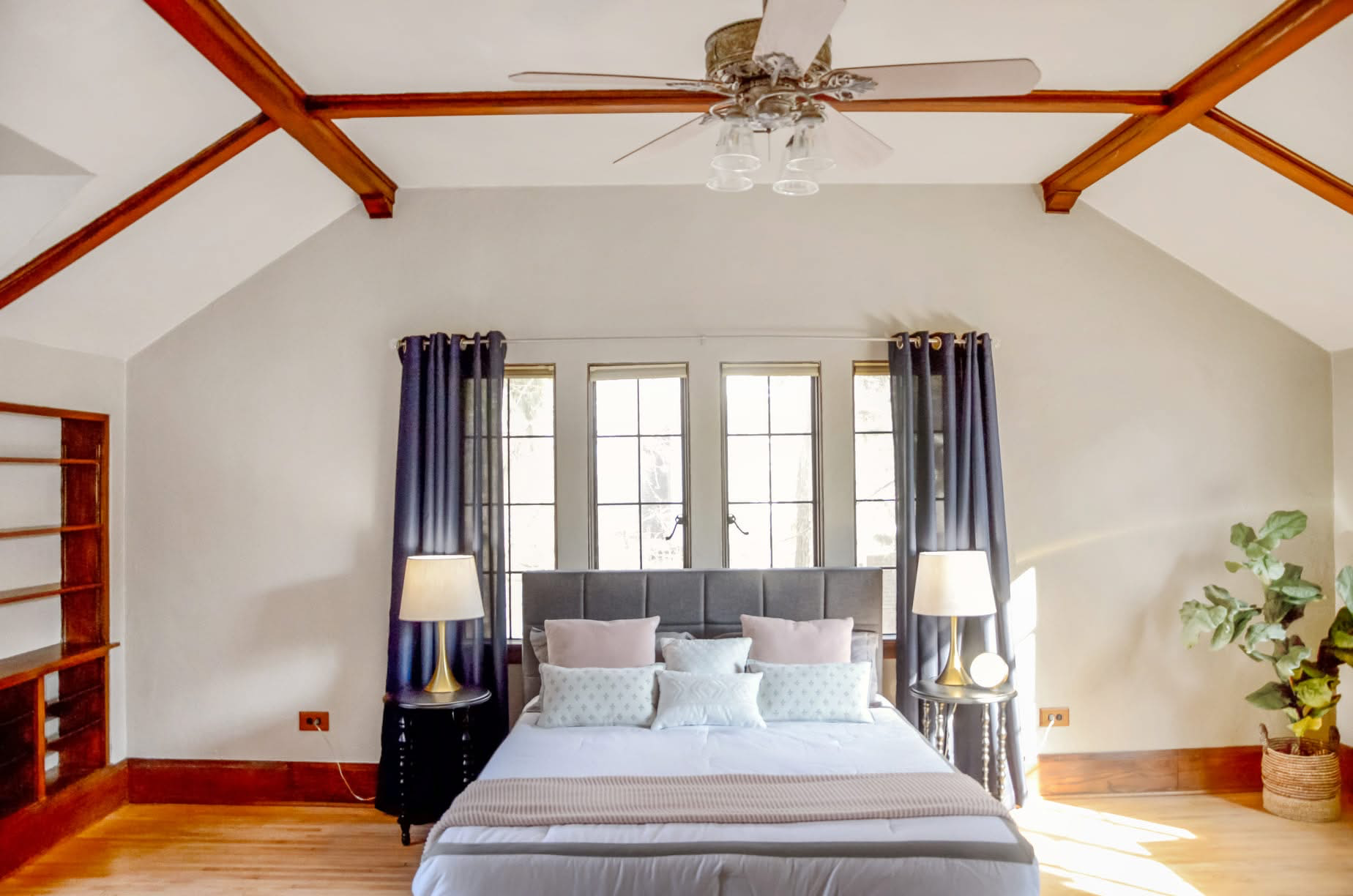 Modern bedroom with white walls, wooden floor, and vaulted ceiling with exposed wooden beams. The bed has a padded headboard, multiple pillows, and a beige blanket. Flanking the bed are black side tables with lamps. There are three windows behind the bed with dark blue curtains, and a potted plant is on the right side of the room.