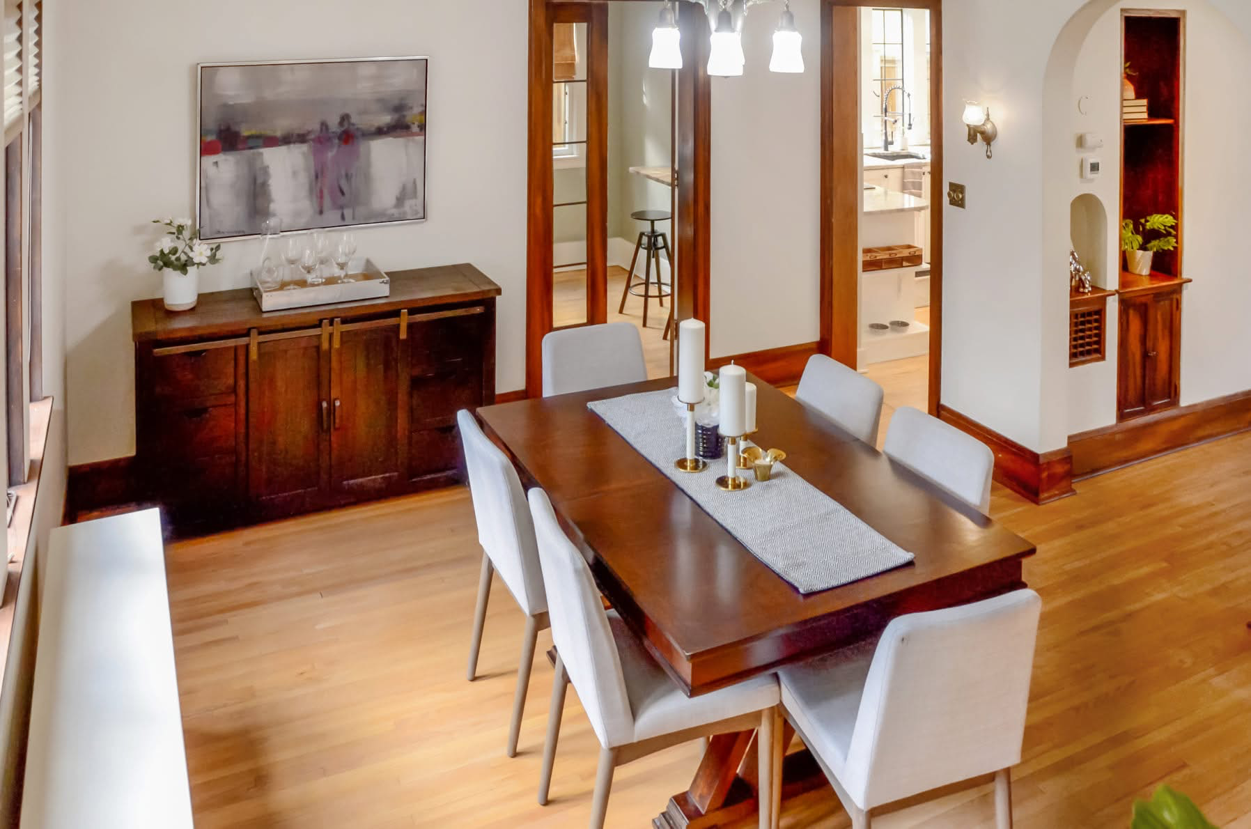A dining room with a wooden table and six light-colored chairs, a wooden sideboard with a tray and wine glasses, a large abstract painting on the wall, a chandelier above the table, and a doorway leading to a kitchen with a barstool. There is also a small built-in shelf with a plant and decorative items.