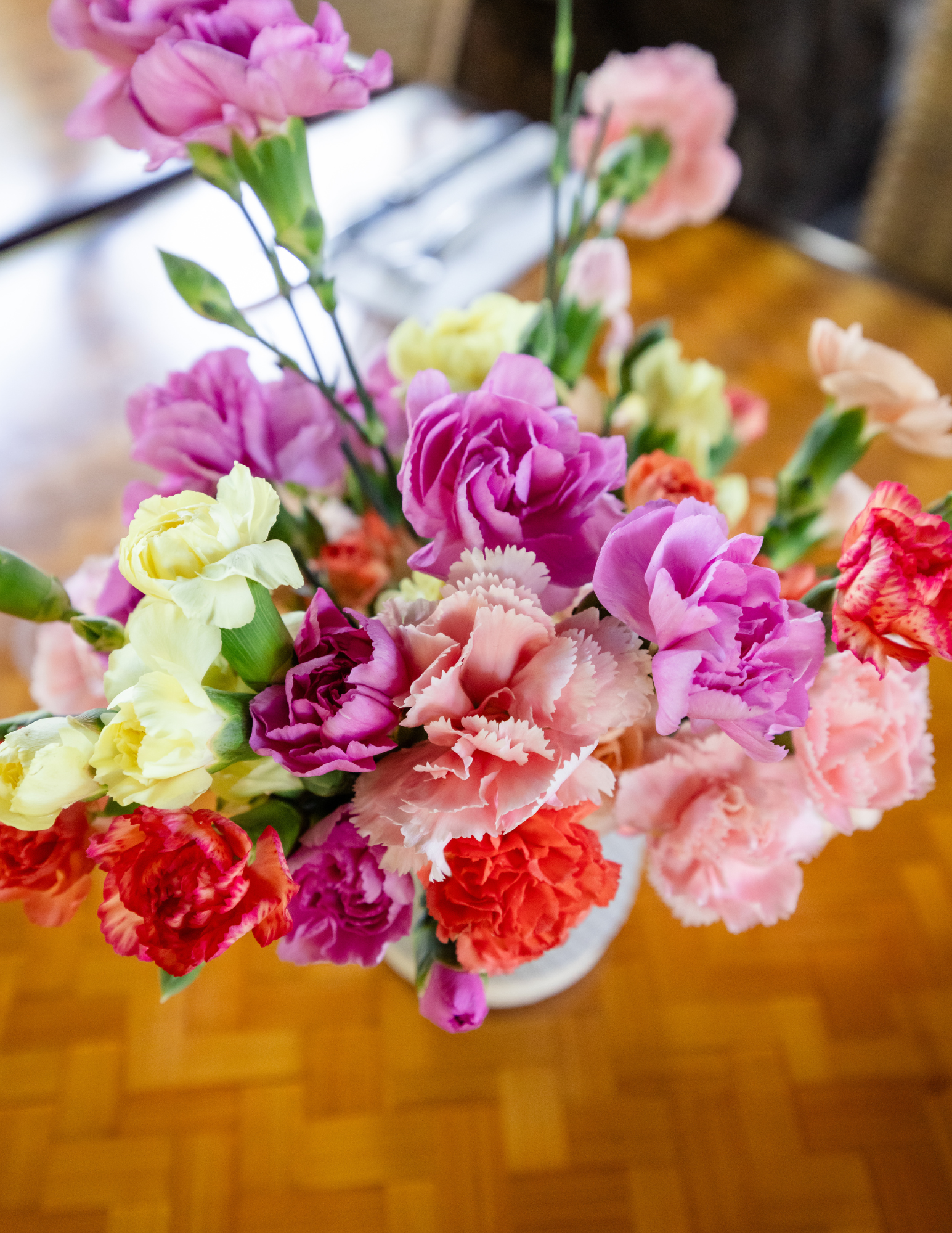 Close-up of a colorful bouquet of carnations in shades of pink, red, peach, and yellow arranged in a small vase on a wooden table.