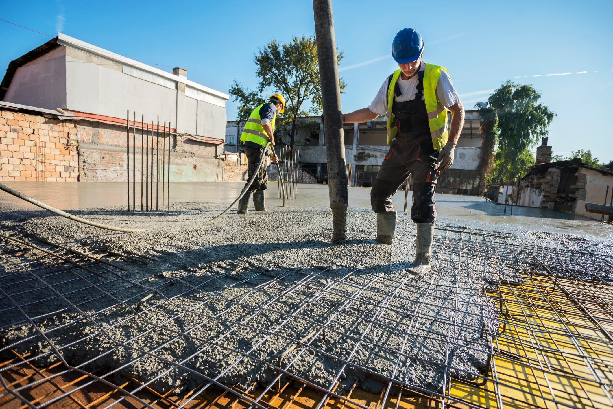 Two construction workers wearing safety vests and helmets are pouring and leveling concrete on a building site during daytime. Rebar is visible in the concrete, with residential buildings and trees in the background.