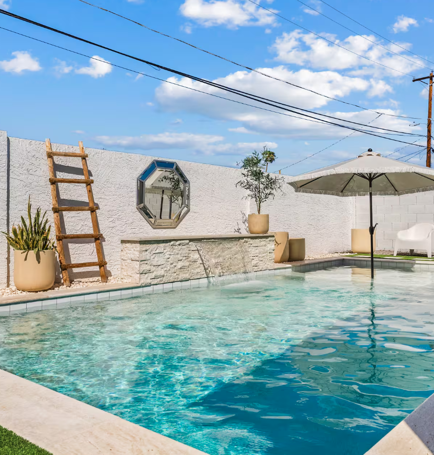 Backyard pool area with a white fence, potted plants, a water feature with a small cascade, a large umbrella, a lounge chair, and a white outdoor chair under a sunny sky.
