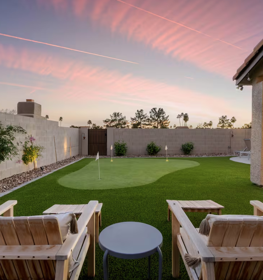 Backyard with a mini golf putting green, two wooden chairs with cushions, a small round table, a concrete block wall, and palm trees in the background under a pink and blue sky at sunset.