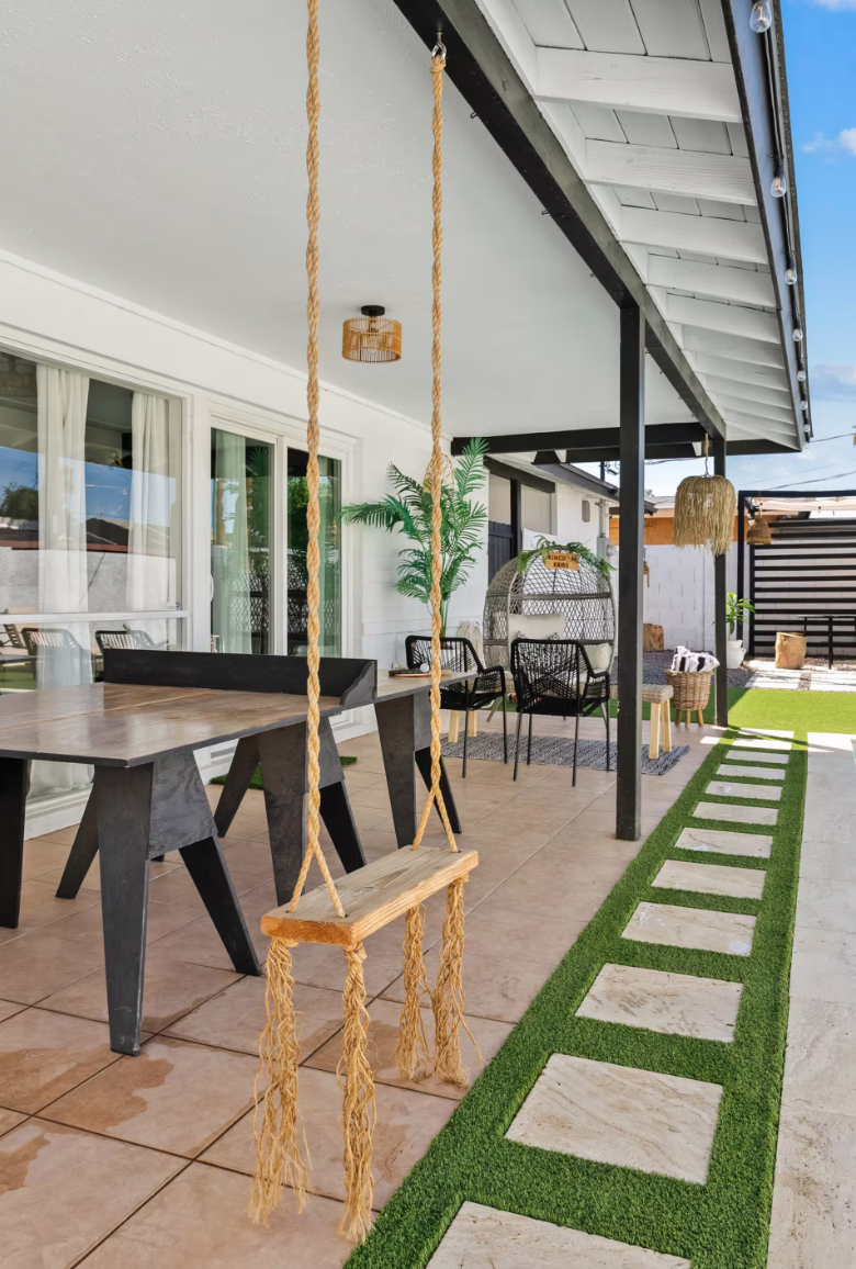 Outdoor patio with a wooden swing hanging from the ceiling, outdoor dining table, black chairs, potted plant, shaded area, and decorative pathway with grass and stone tiles.