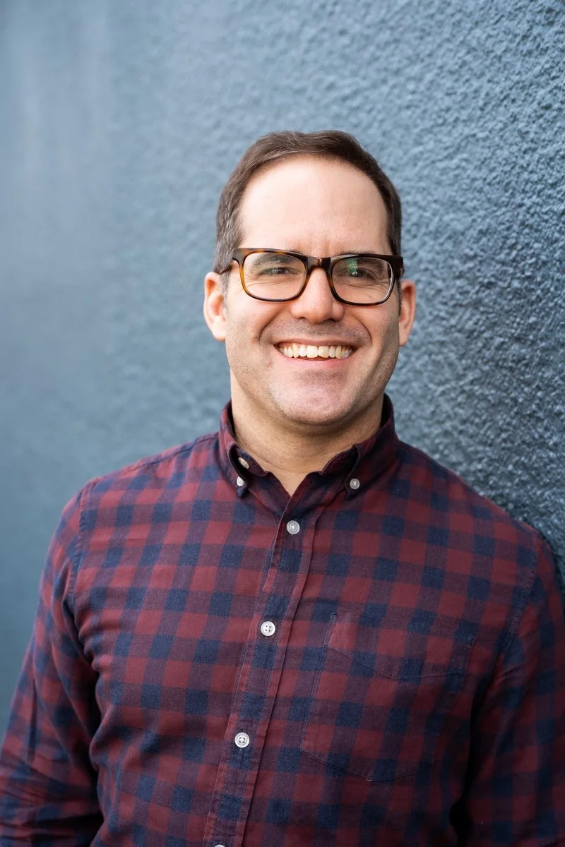 A smiling man with glasses wearing a red and blue checkered shirt standing against a dark textured wall.
