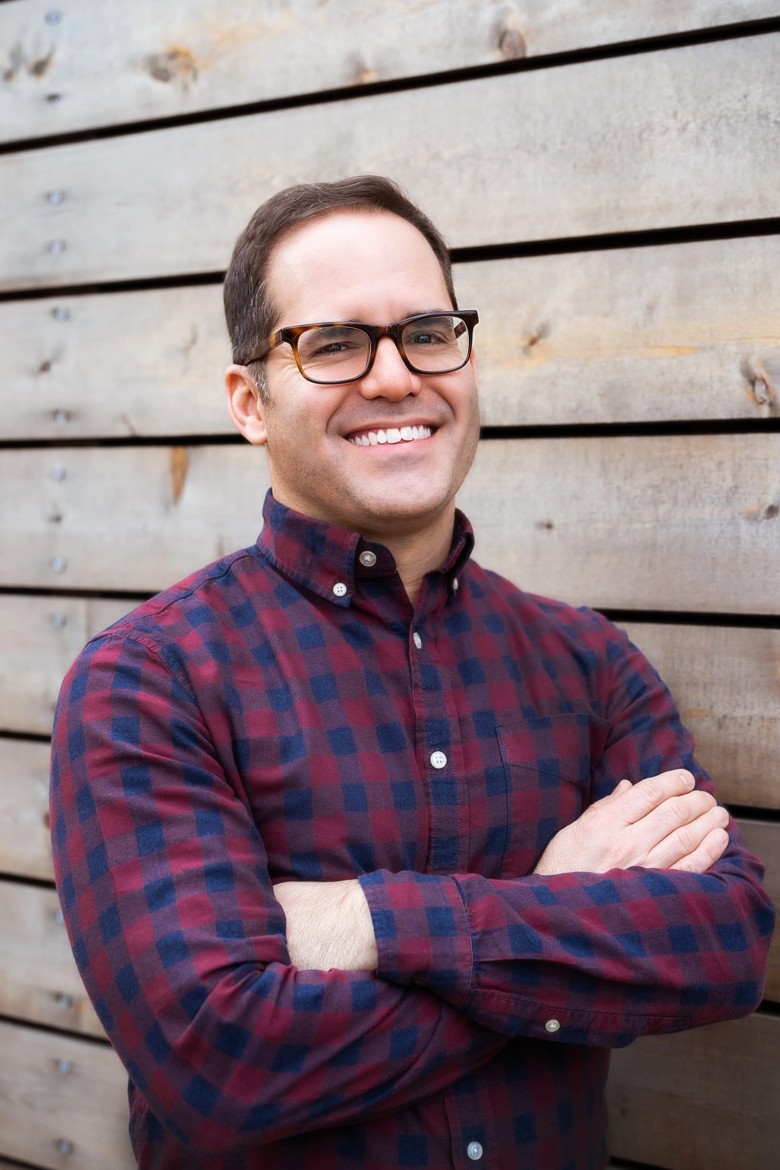 A smiling man with glasses, wearing a red and blue checkered shirt, standing with arms crossed in front of a wooden wall.