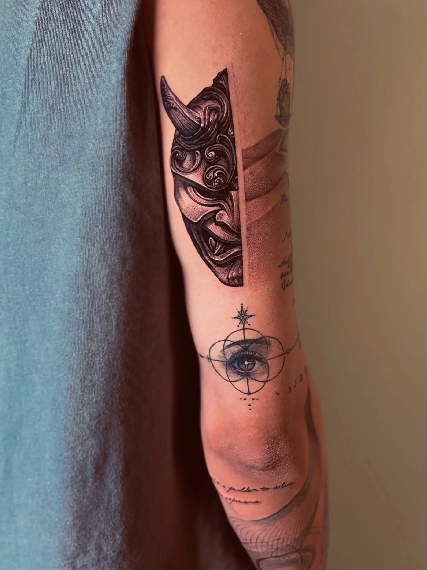 Tattoo of a detailed, stylized demon mask on upper arm, with additional geometric and eye tattoos on lower arm.