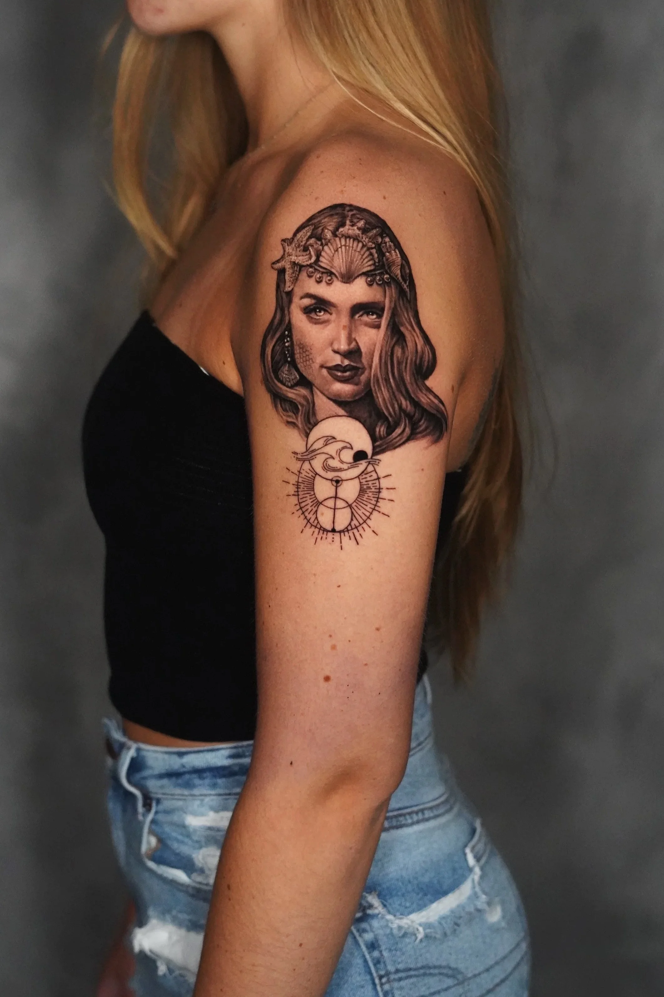 Tattoo of a woman's face with wavy hair, wearing a seashell headpiece, above a design of a mermaid with long flowing hair, a fish tail, and a circular geometric pattern.